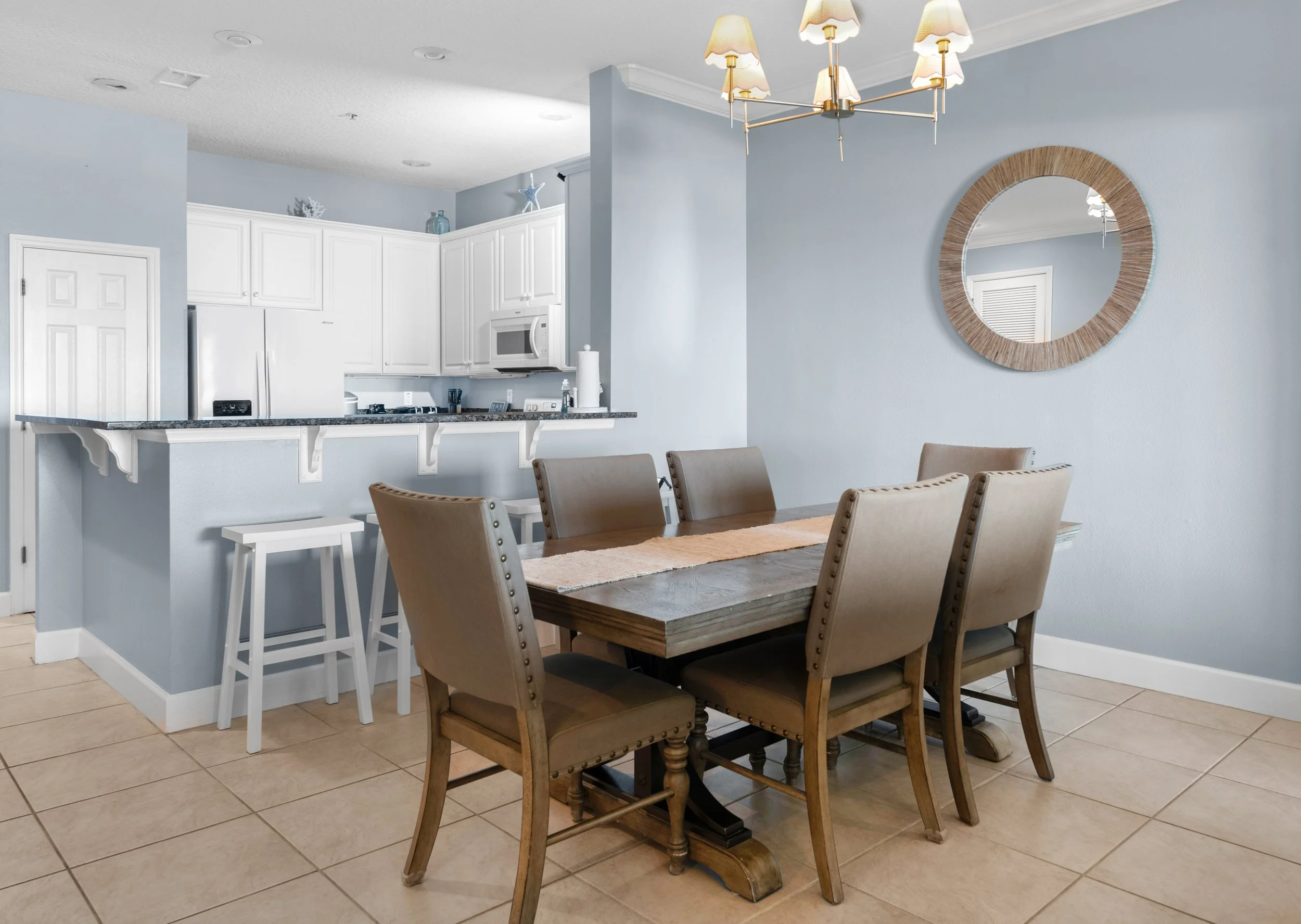 Dining room with six beige upholstered chairs around a wooden table and a light blue wall with a round mirror. A chandelier hangs above the table. In the background, a kitchen with white cabinets, a microwave, and a refrigerator.