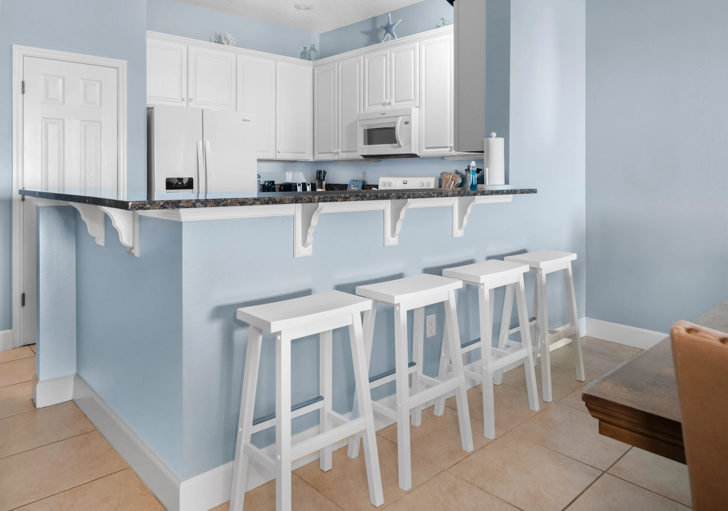 Modern kitchen with white cabinets, a granite countertop, blue walls, and four white bar stools at a breakfast bar.