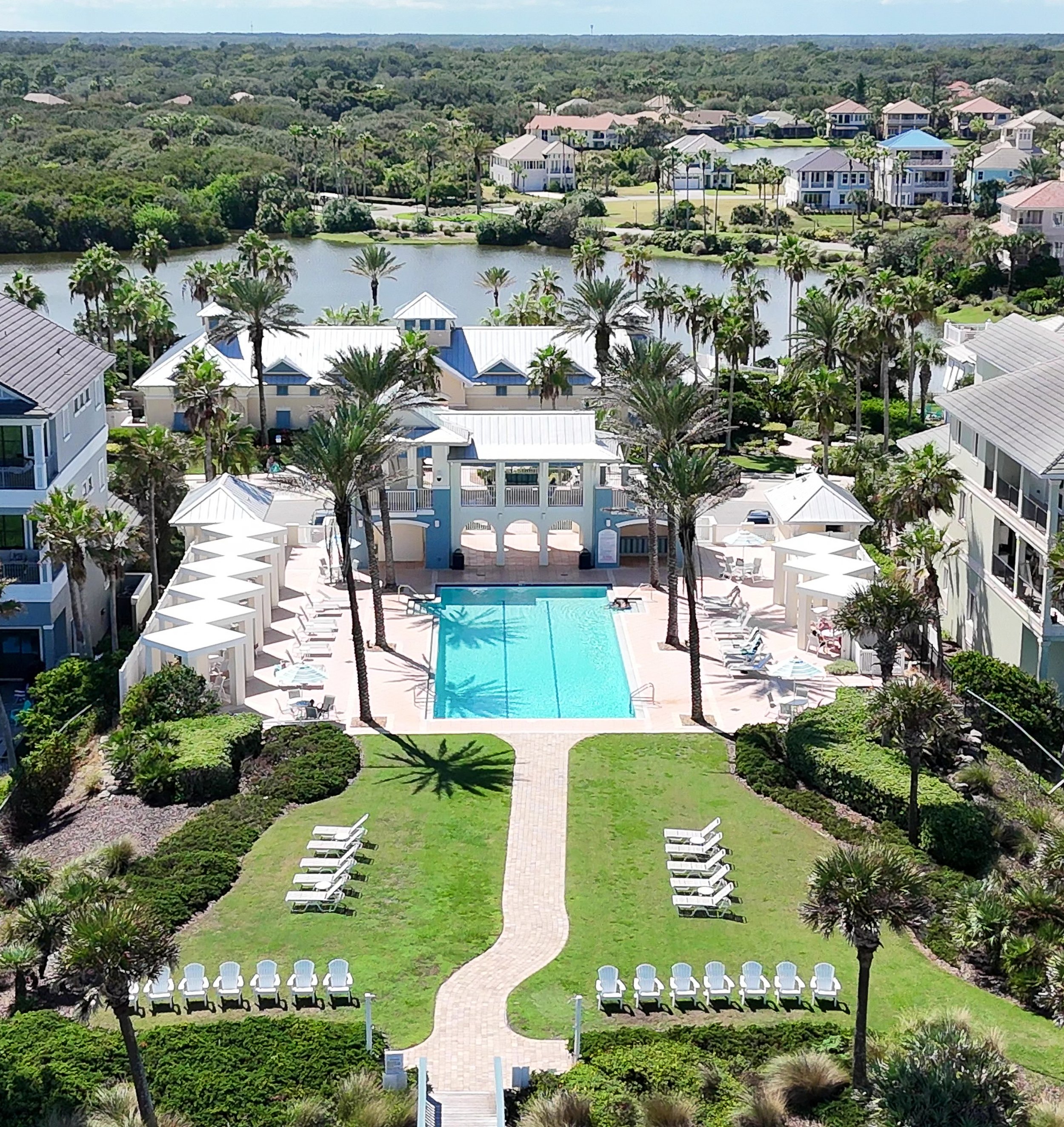 An aerial view of a resort-style pool area with a rectangular swimming pool surrounded by palm trees, lounge chairs, and shaded cabanas. There are multi-story buildings on either side and a lush green landscape with a pond in the background.