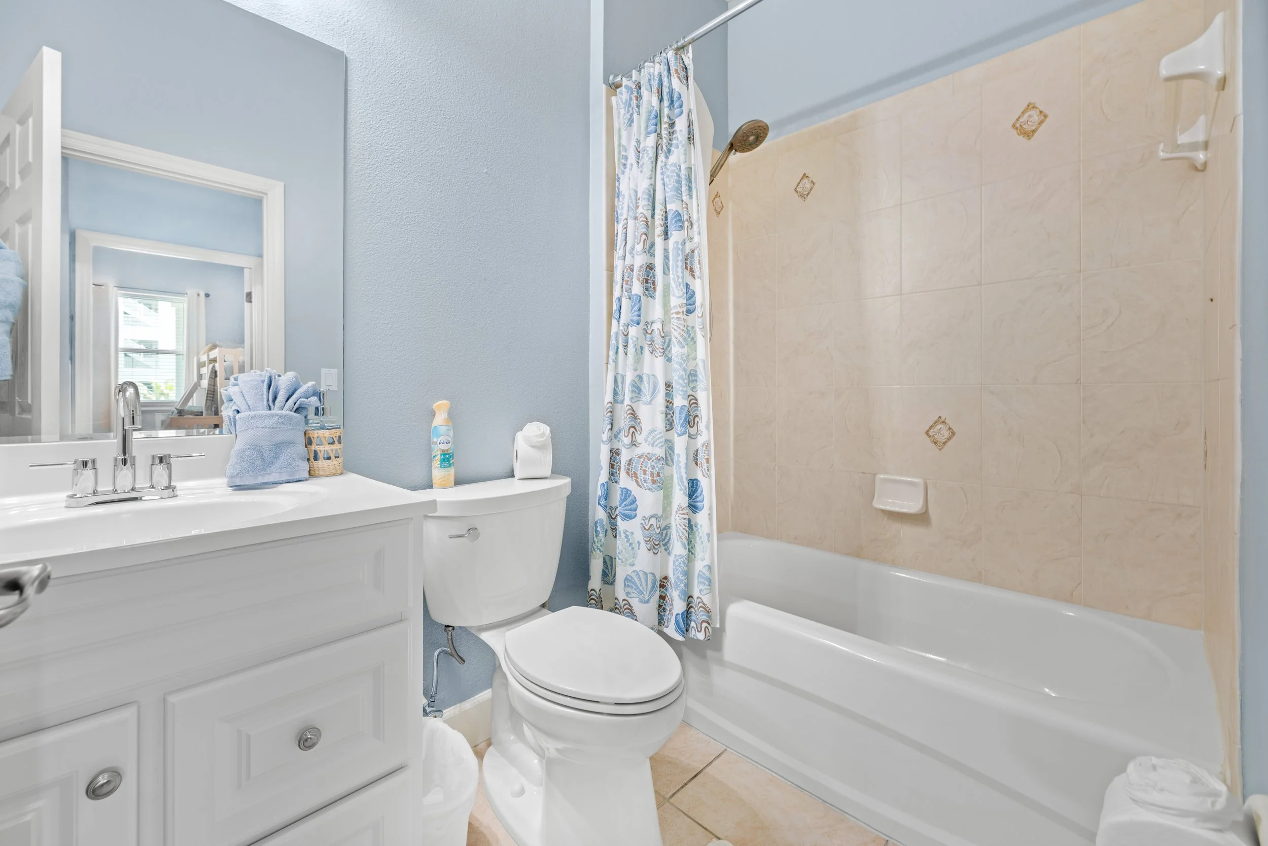 Bathroom with white vanity, mirror, toilet, and shower with beige tiled wall and a white bath tub, blue walls, and a colorful shell-patterned shower curtain.