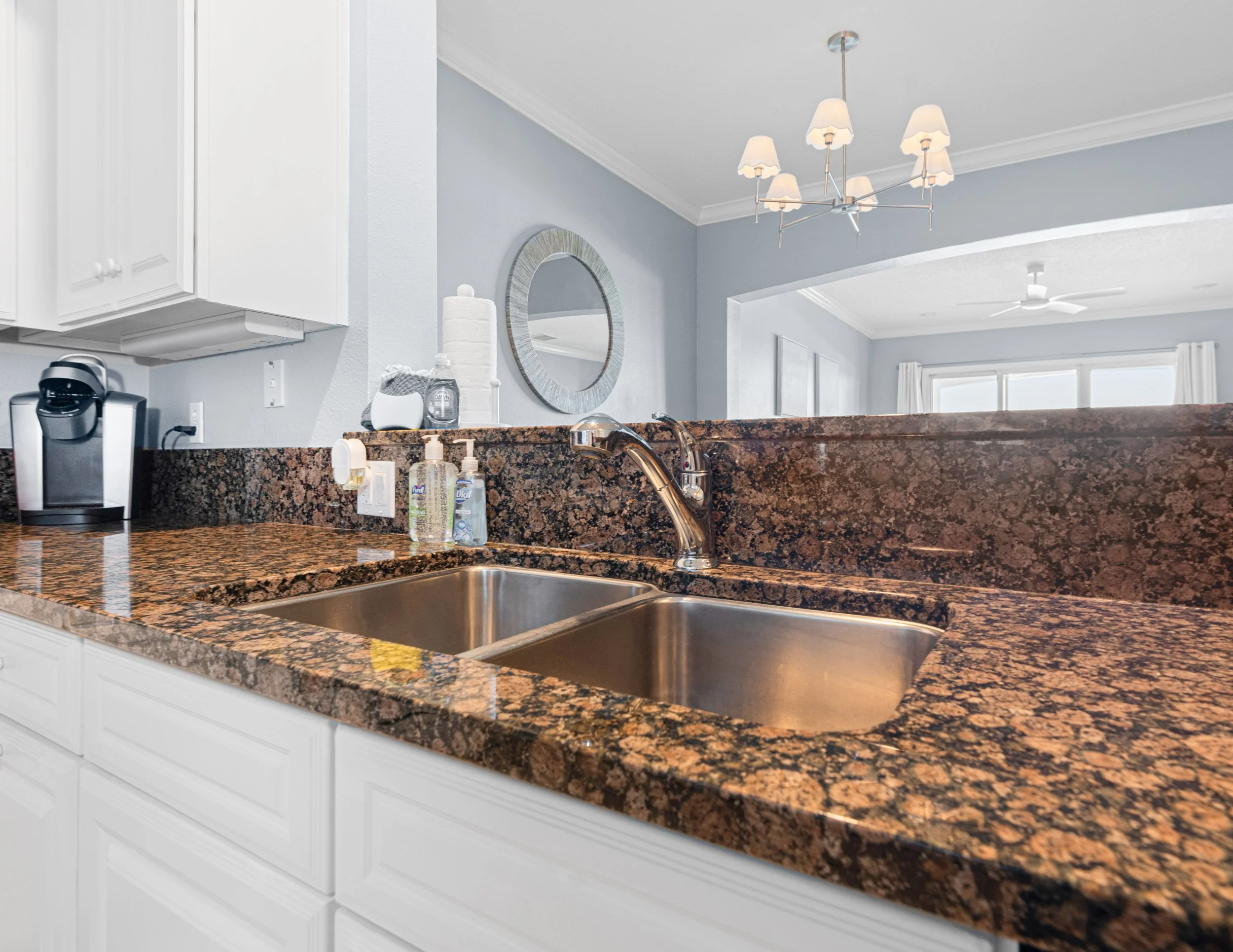 Kitchen countertop with double sink, soap dispensers, a coffee machine, and white cabinets, opening to a living area with a chandelier and ceiling fan.