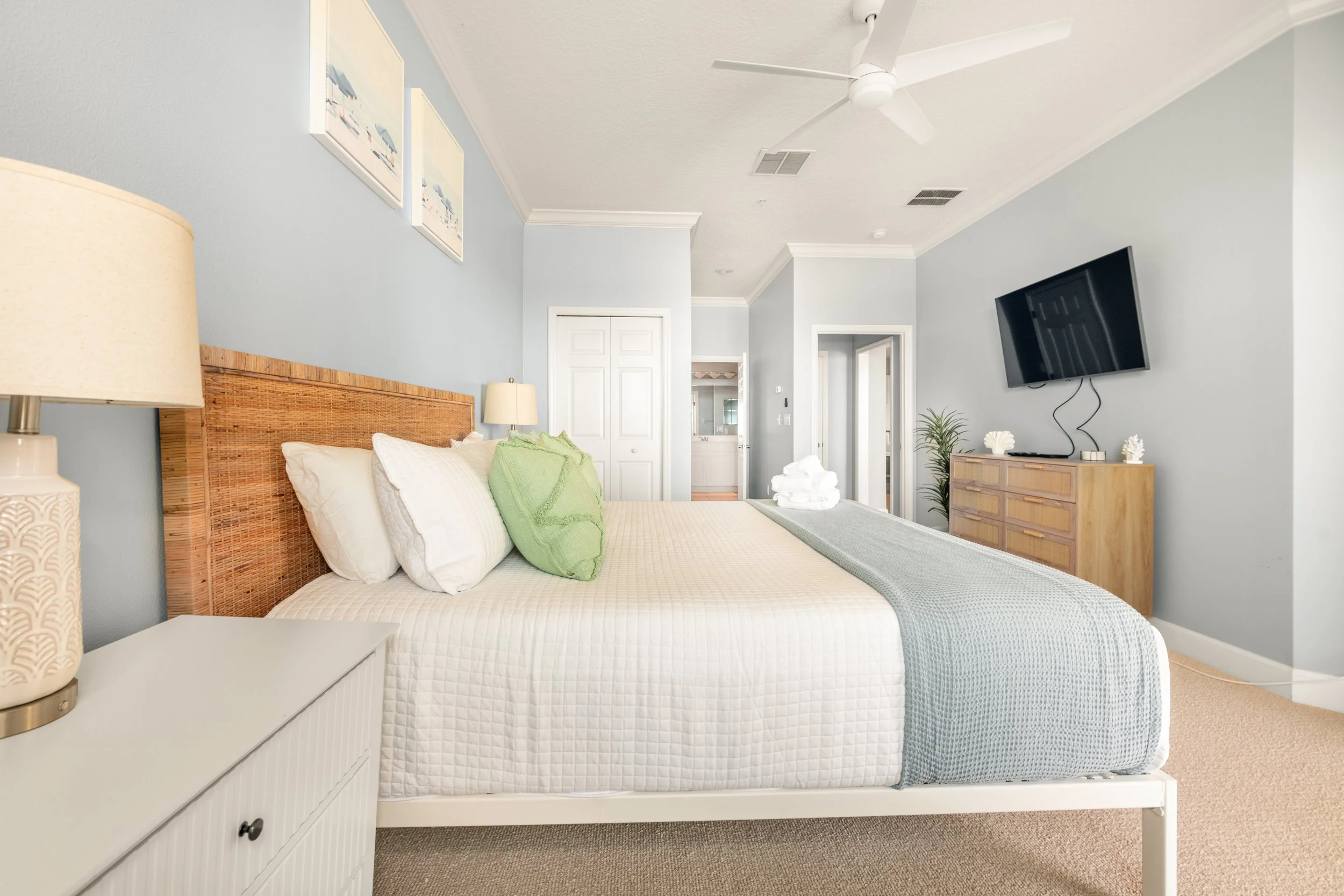A well-made hotel bedroom with a bed, pillows, lamp, wall art, dresser, TV, ceiling fan, and a plant.