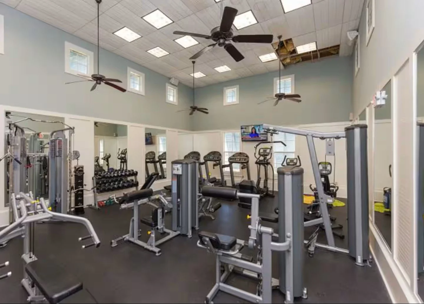 A gym with various weight machines and cardio equipment, high ceilings with ceiling fans, and a television mounted on the wall.