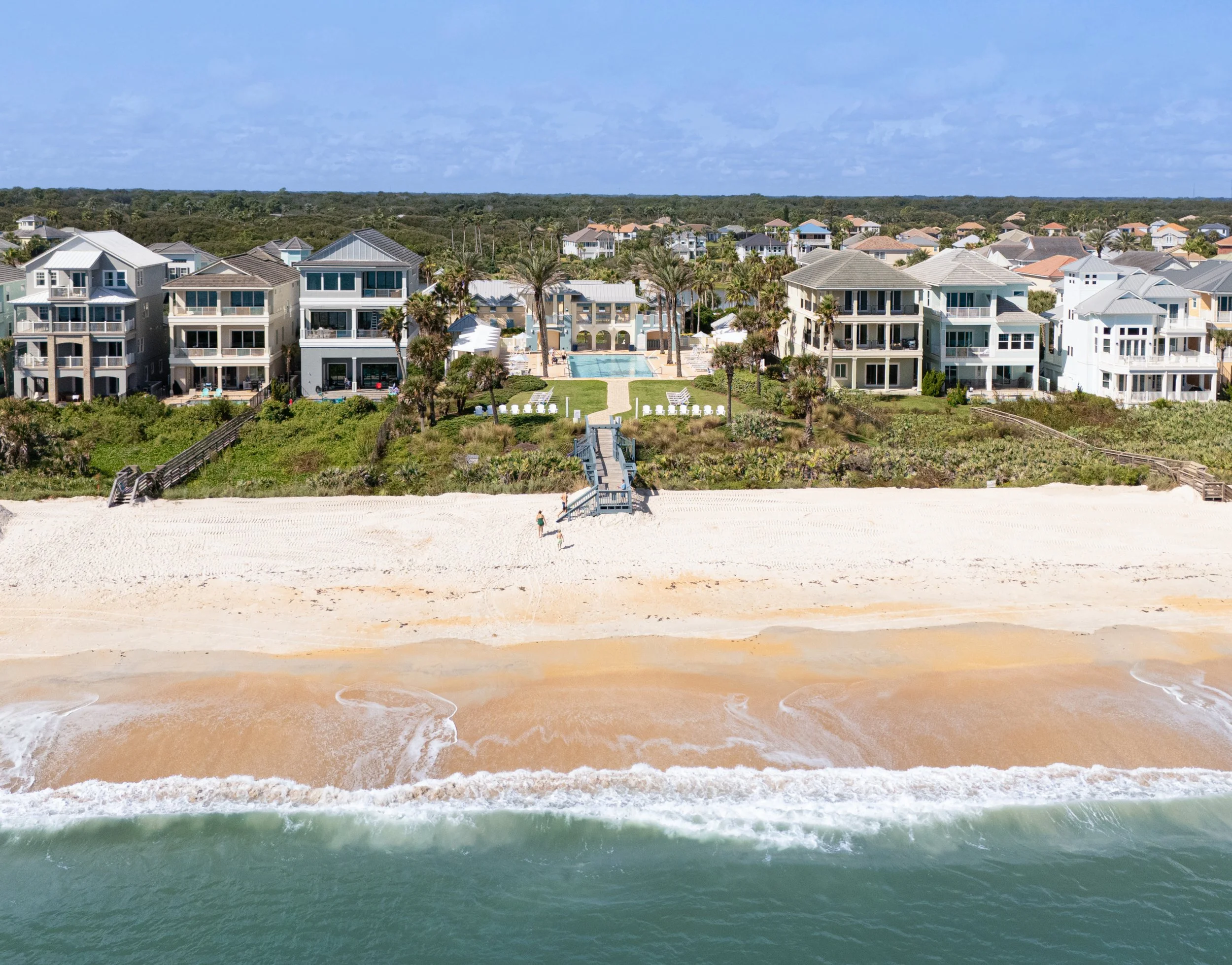 Aerial view of beachfront houses with a swimming pool, palm trees, and a sandy beach with waves in the foreground.