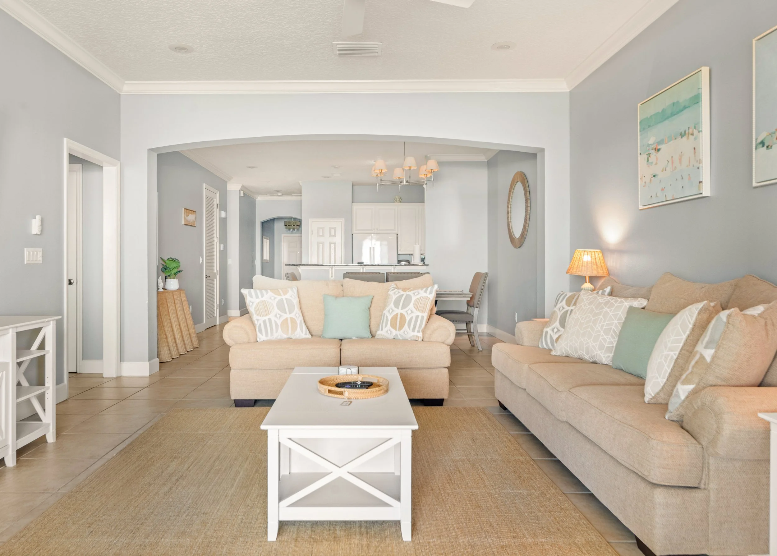 Living room with beige sofas, pillows, a white coffee table, and wall art, leading to an open kitchen area.