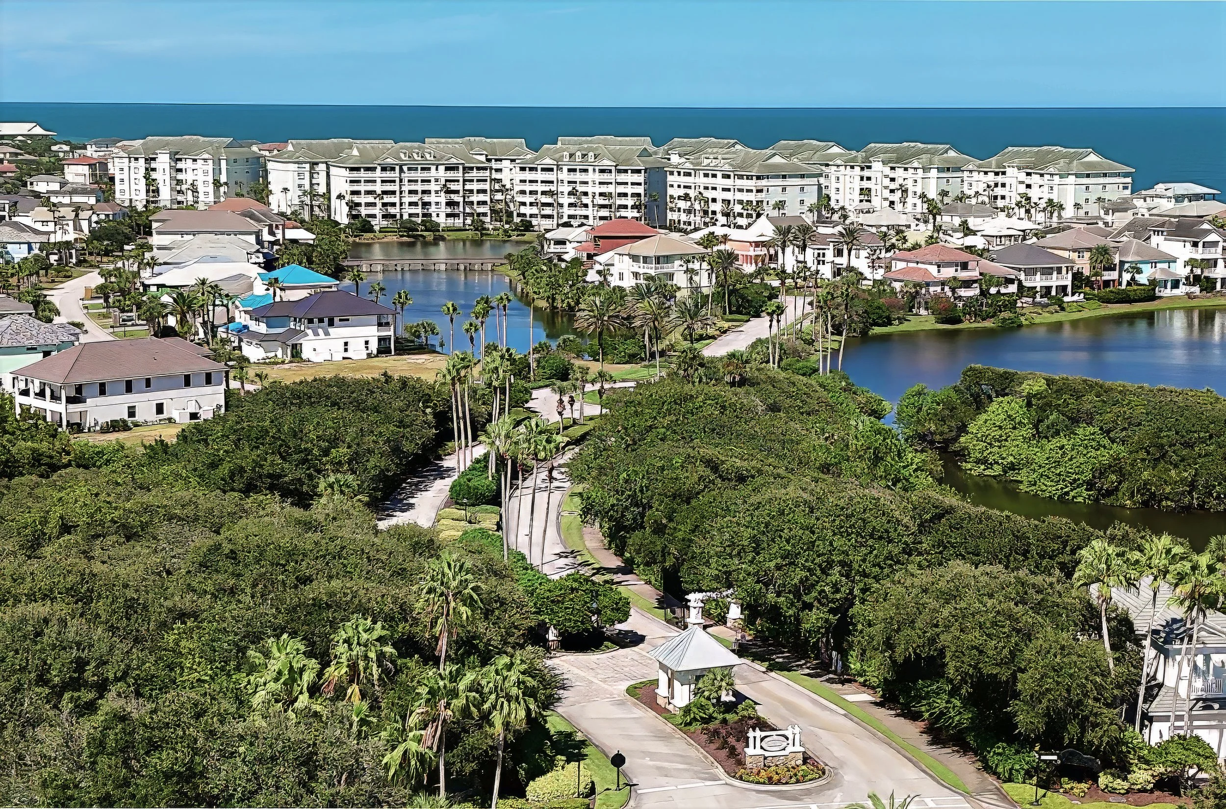 Aerial view of a resort with white buildings, small lakes, lush green trees, and a clear blue ocean in the background.