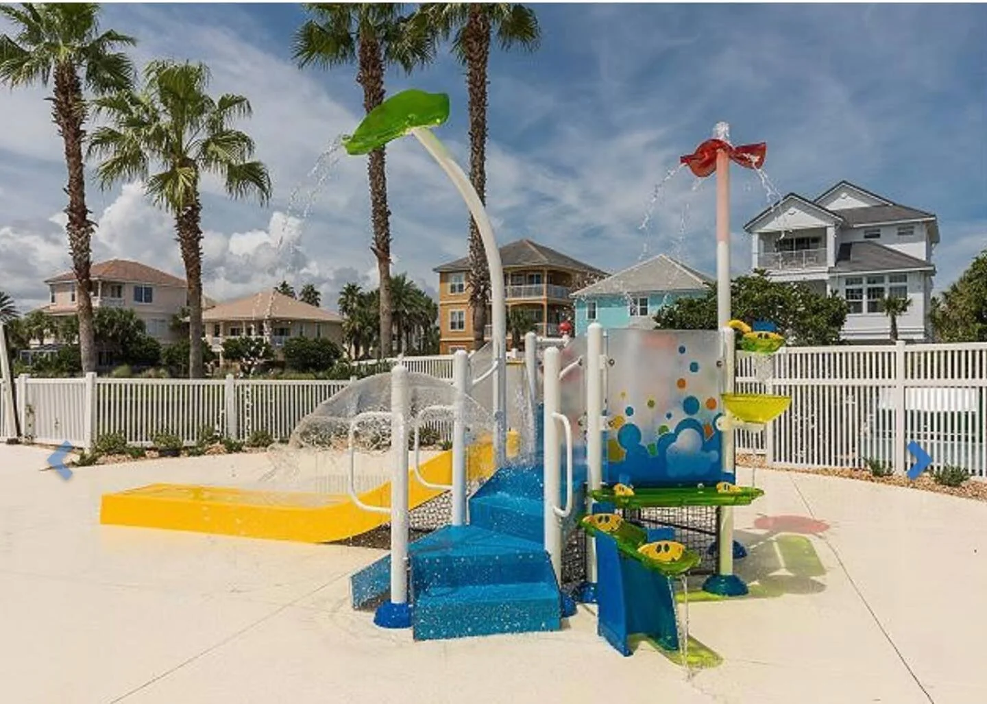 Colorful water playground with slides, water sprayers, and fountains, set against residential houses and palm trees on a sunny day.