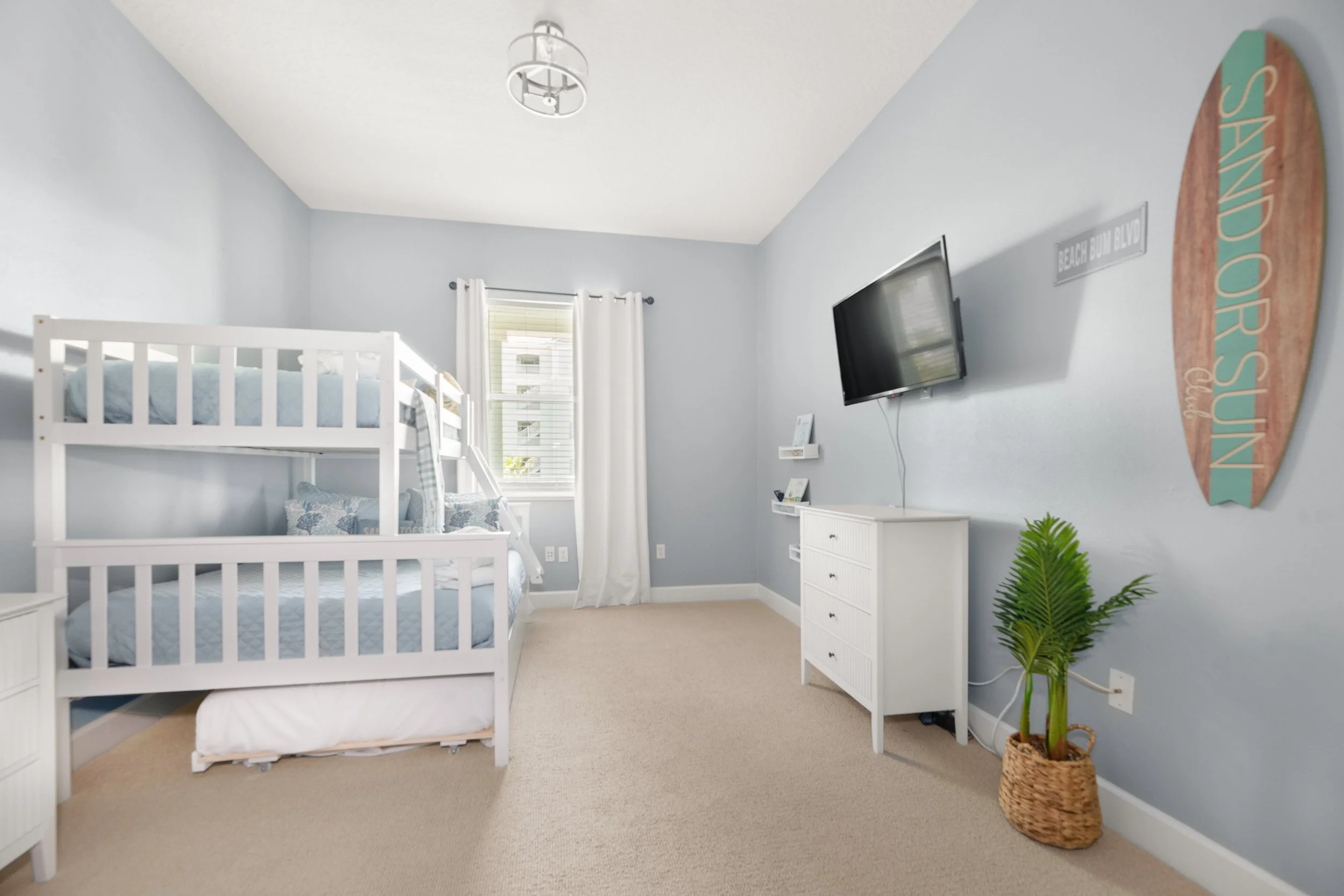 Bright bedroom with light blue walls, white bunk beds with blue bedding, a window with white curtains, a wall-mounted TV, a white dresser, and a potted plant in a woven basket.