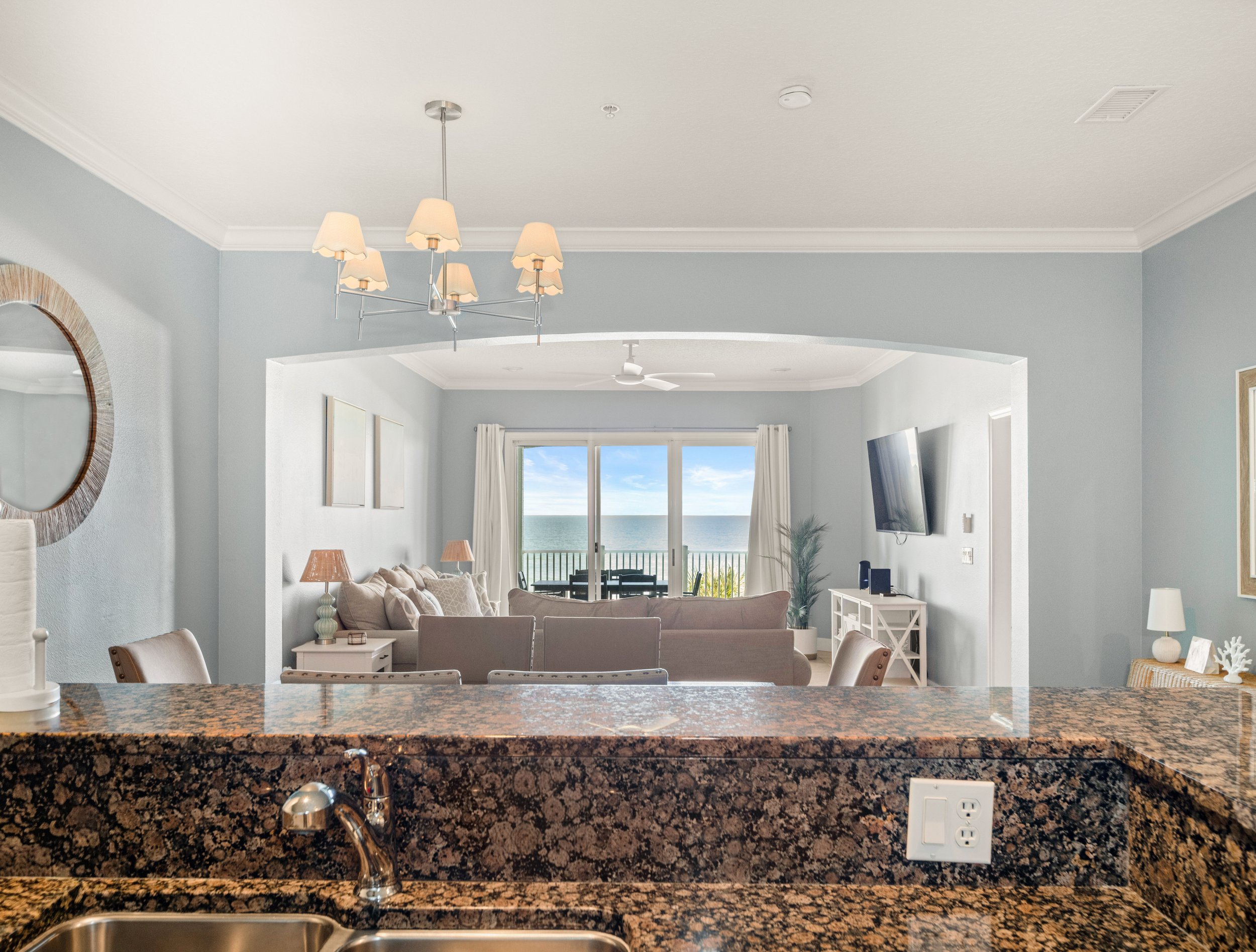 Living room with ocean view through sliding glass doors, furnished with a sofa, armchairs, and a wall-mounted TV, viewed from a kitchen with a granite countertop and sink.