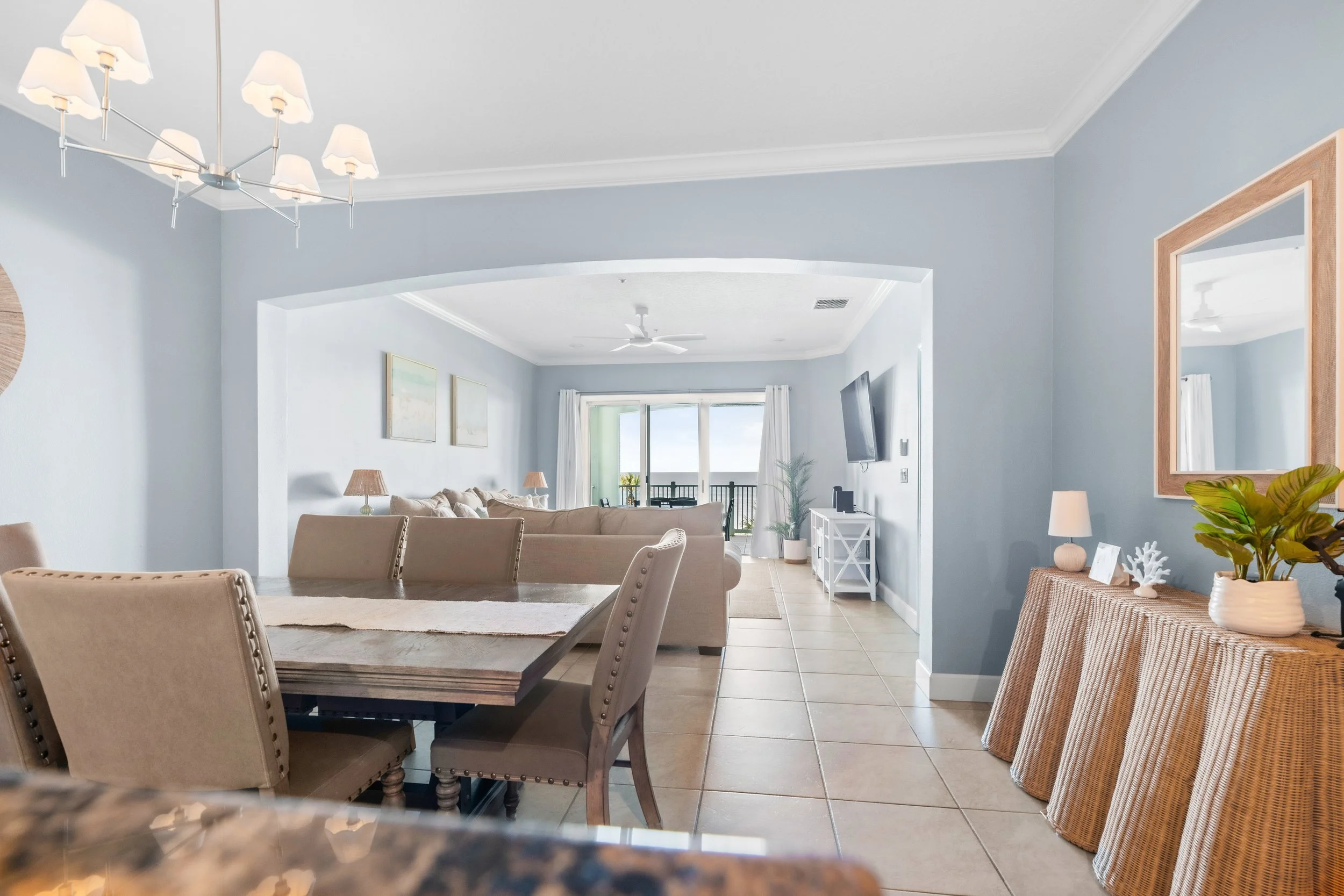 Living room and dining area in a beachside condo with light blue walls, tiled floors, a dining table with beige chairs, a sofa, a wall-mounted TV, and large sliding doors leading to a balcony with an ocean view.