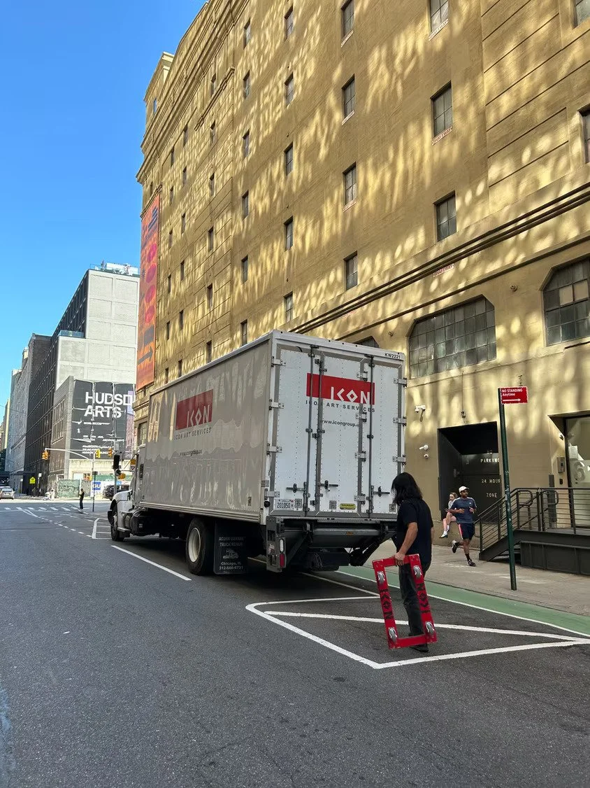 A person stands next to an ICON Art Services white delivery truck parked in an urban street, holding a red moving dolly, with a yellow building and pedestrians in the background.