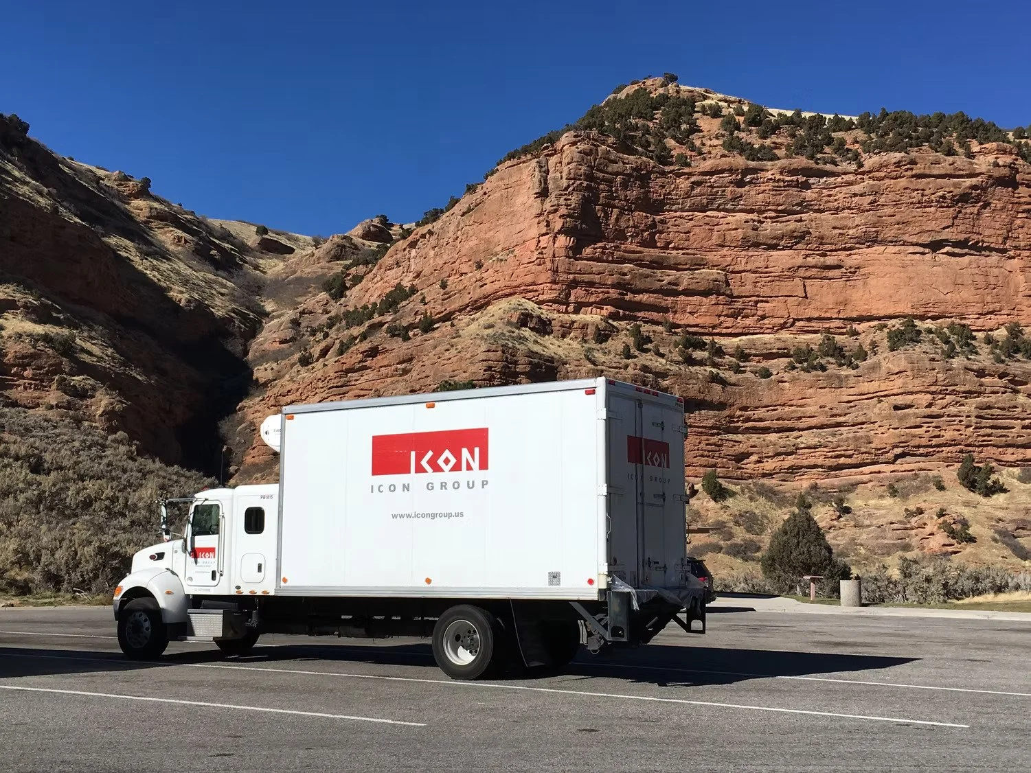 A white delivery truck with ICON Art Services logo on its side, parked on an empty road with a backdrop of red rock formations and a clear blue sky.