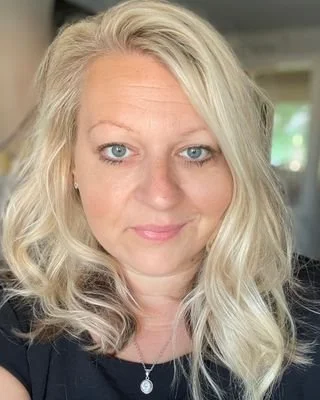 Close-up of a woman with blonde, wavy hair and blue eyes, wearing a black top and a necklace, smiling gently indoors.