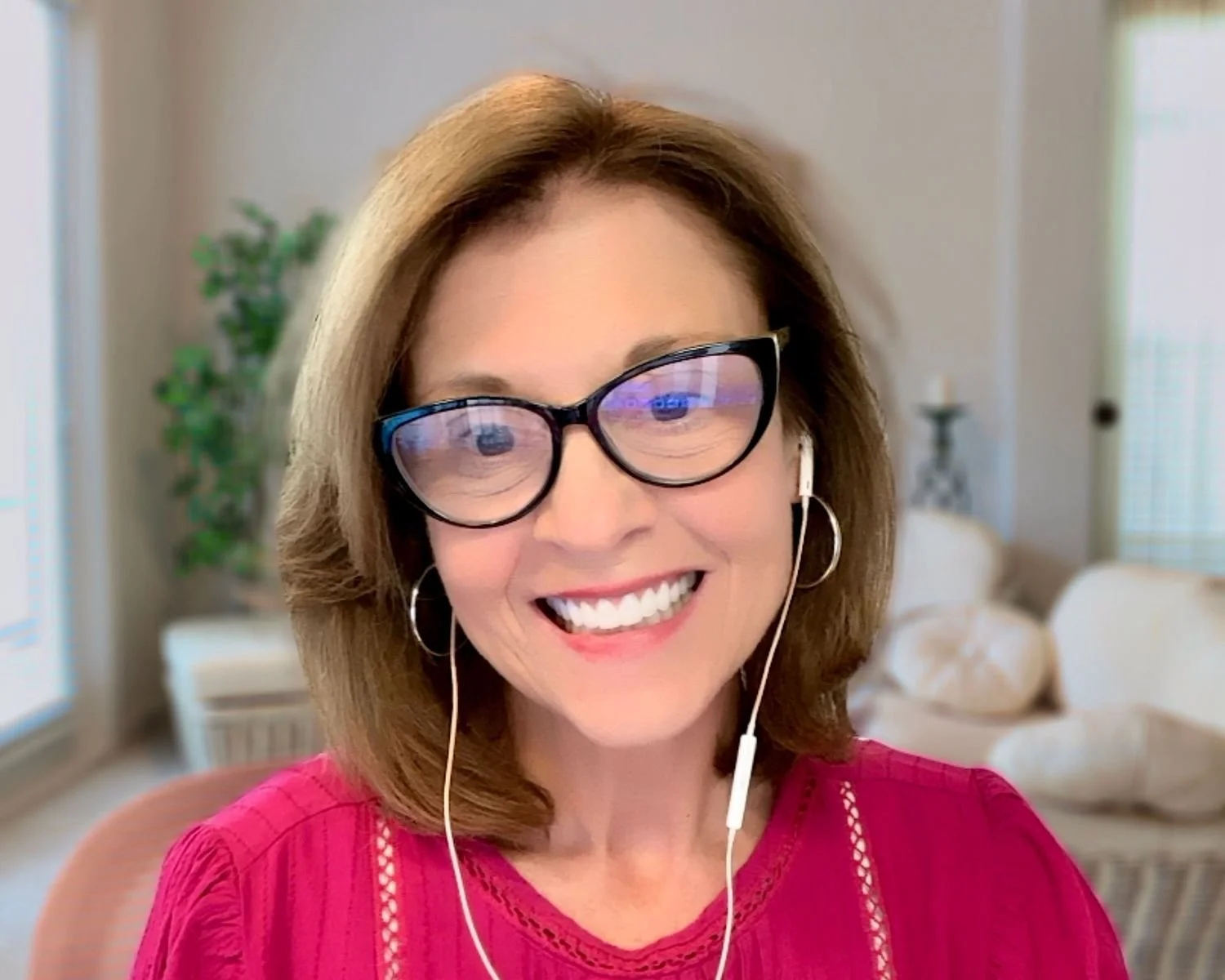 A smiling woman with shoulder-length brown hair and glasses, wearing a pink top, sitting indoors with a background of a living room and a window.