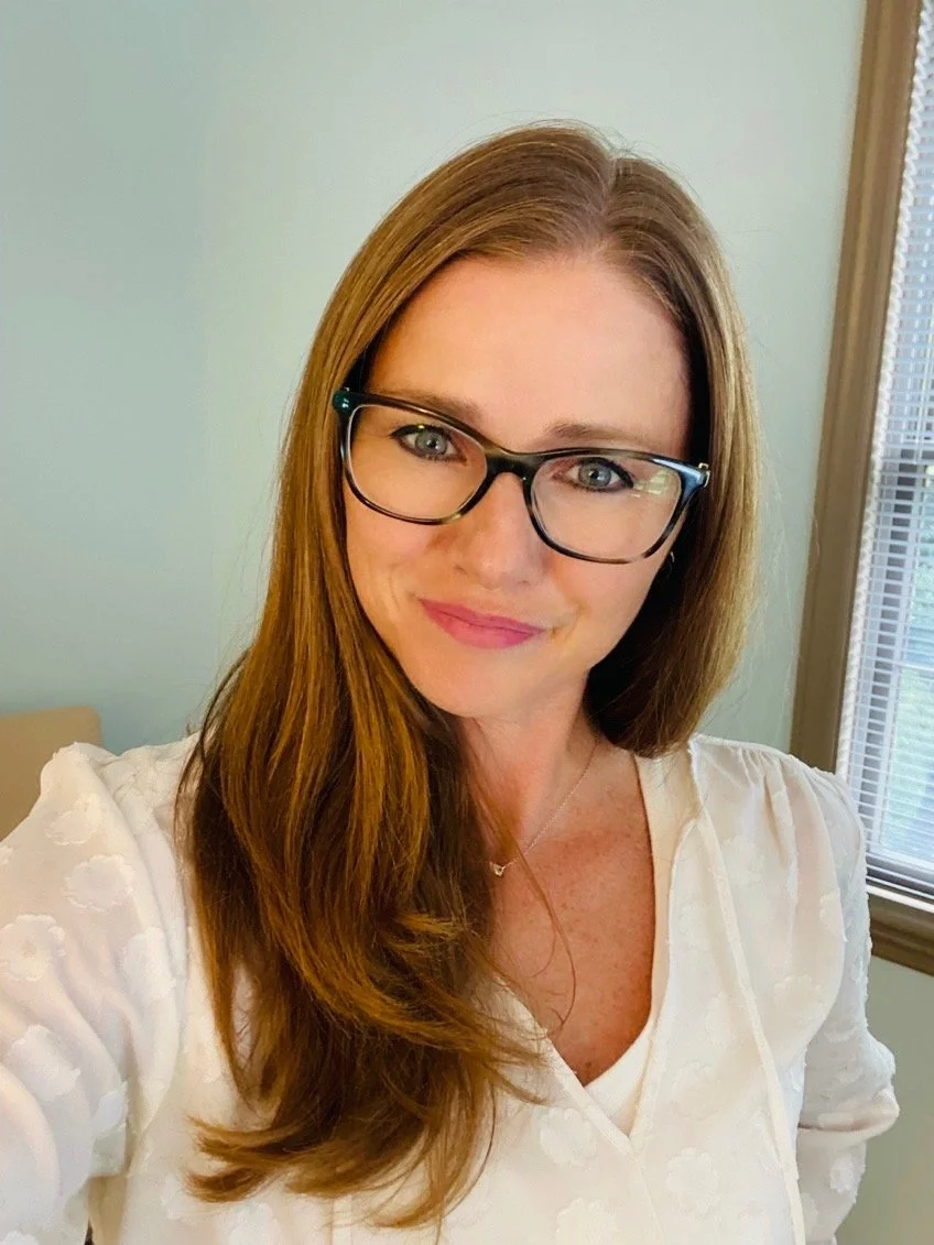 A woman with long brown hair, glasses, and a white embroidered top standing near a window, smiling at the camera.