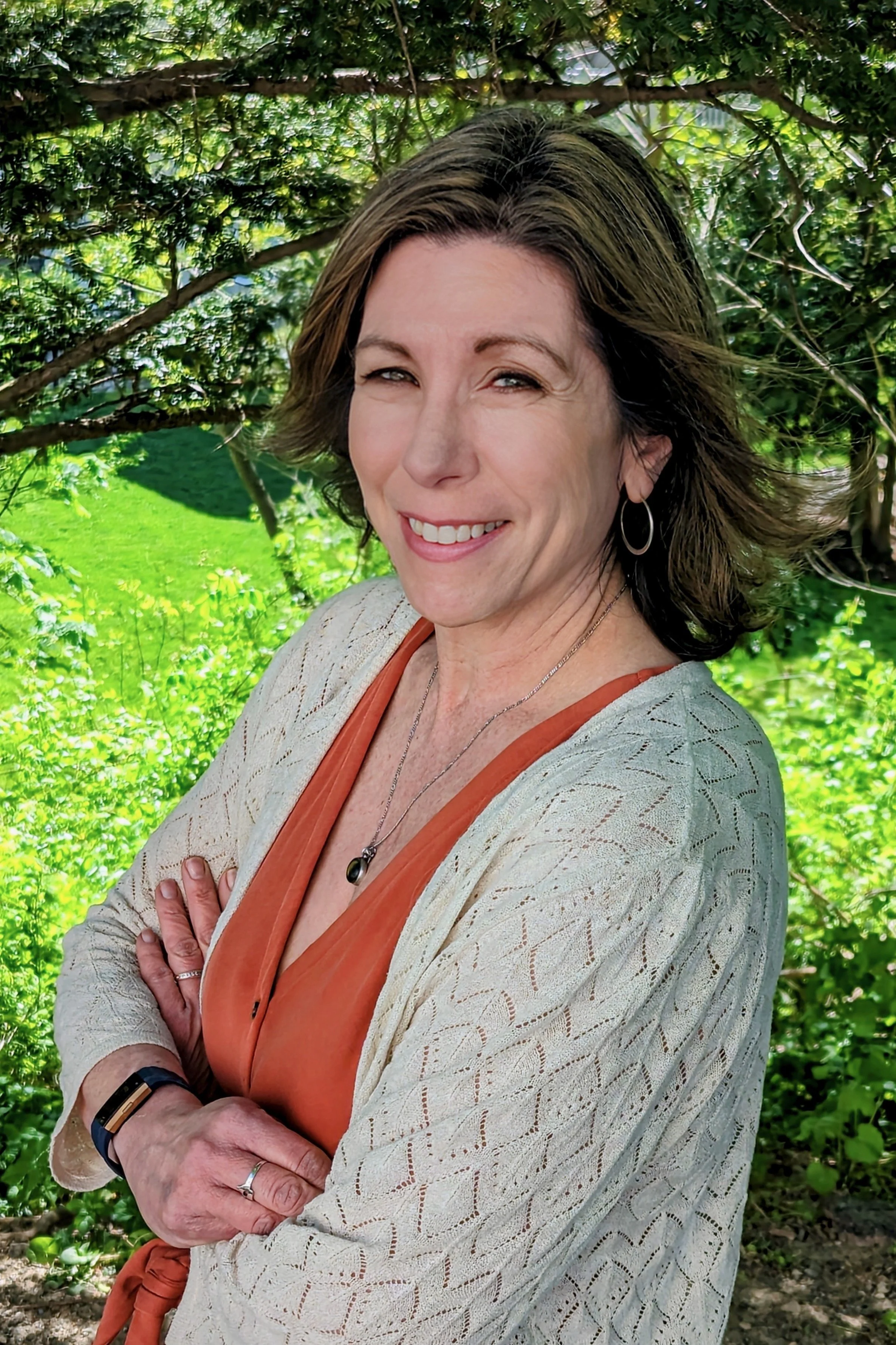 A woman with shoulder-length brown hair smiling outdoors, wearing a beige patterned cardigan over an orange top, with jewelry including earrings, a necklace, and a ring, standing in front of green foliage and a tree branch.