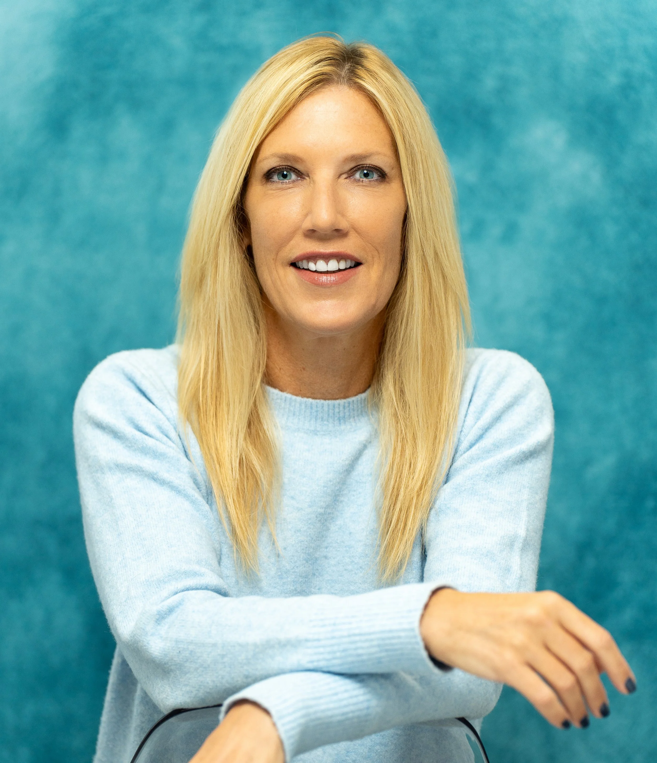 A woman with long blonde hair, blue eyes, and fair skin, wearing a light blue sweater, smiling against a blue background.