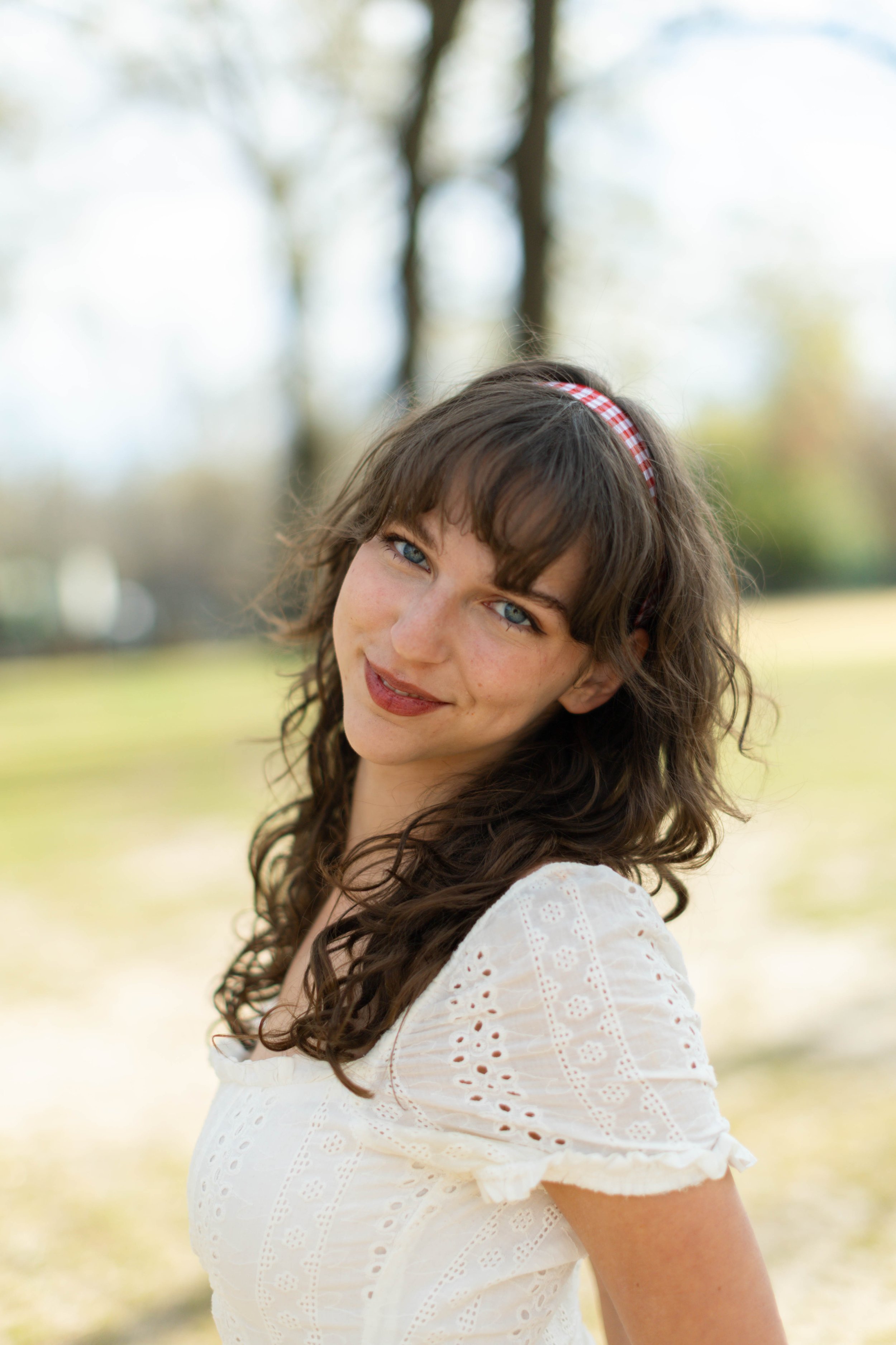 A woman with curly brown hair and blue eyes wearing a white eyelet dress and a red and white checkered headband, standing outdoors with blurred trees in the background.