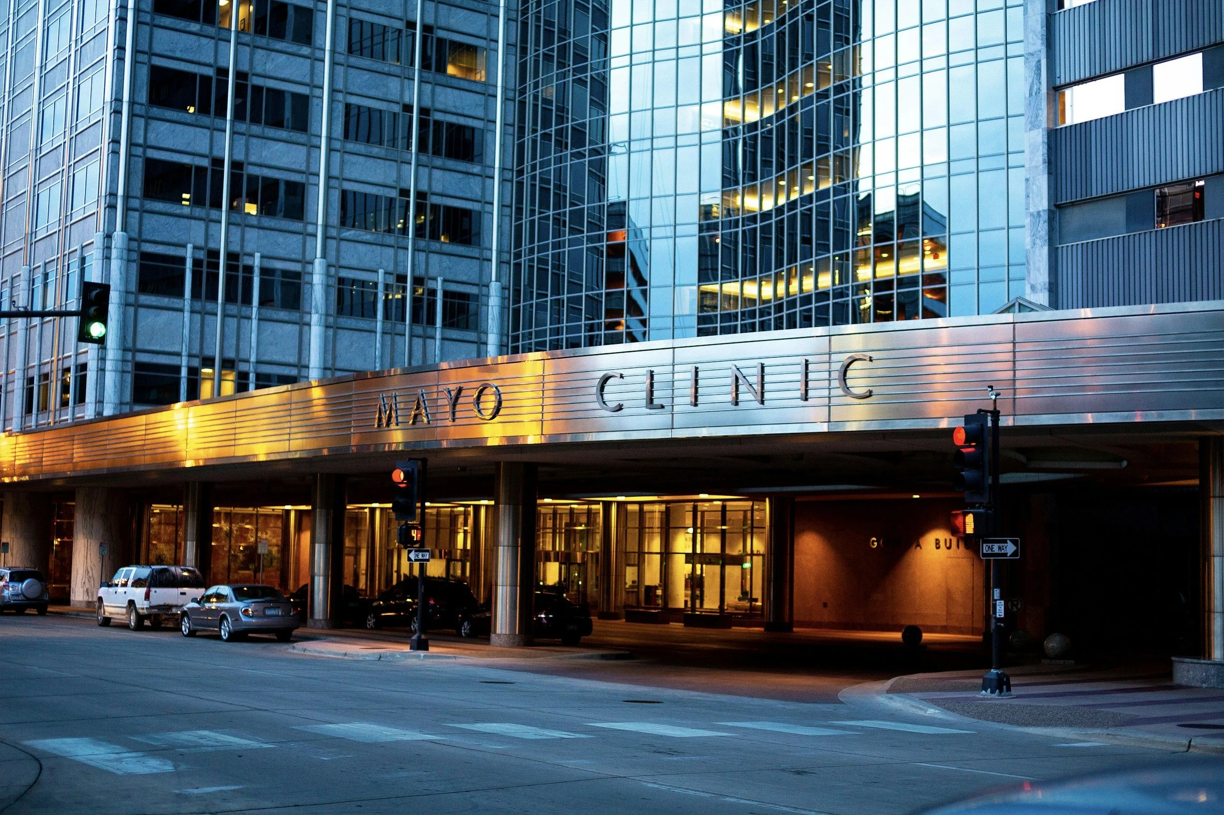 Exterior view of Mayo Clinic building with glass architecture and illuminated entrance at dusk, cars parked nearby, and traffic lights at the intersection