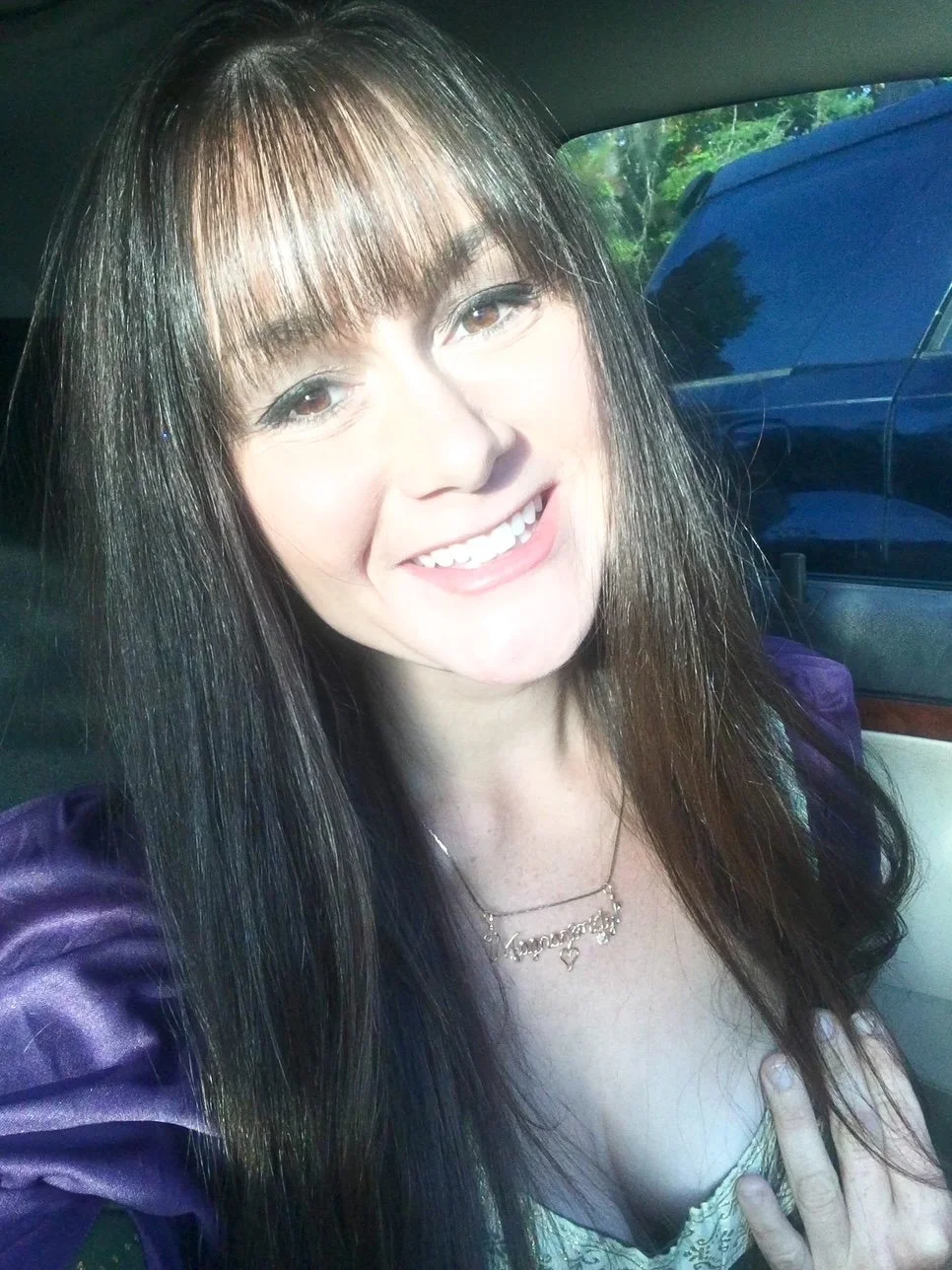 A young woman with long dark hair and bangs, smiling and showing a peace sign, sitting in a car with sunlight shining on her face.