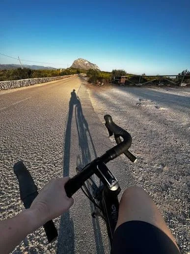 A view of bike handlebars and a long shadow of the cyclist projected on a paved road with a mountain in the background