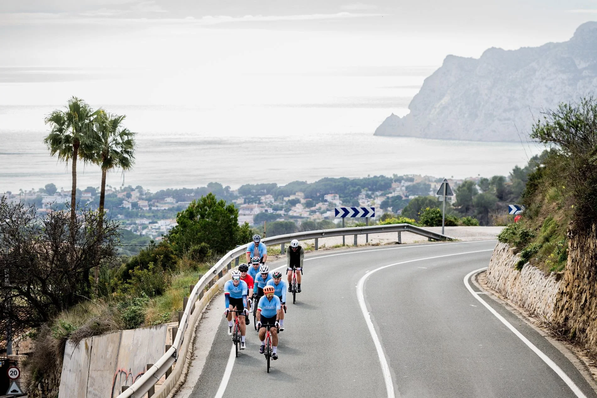 Eight bike riders ascending a paved and curvy road with the sea and mountains in the background