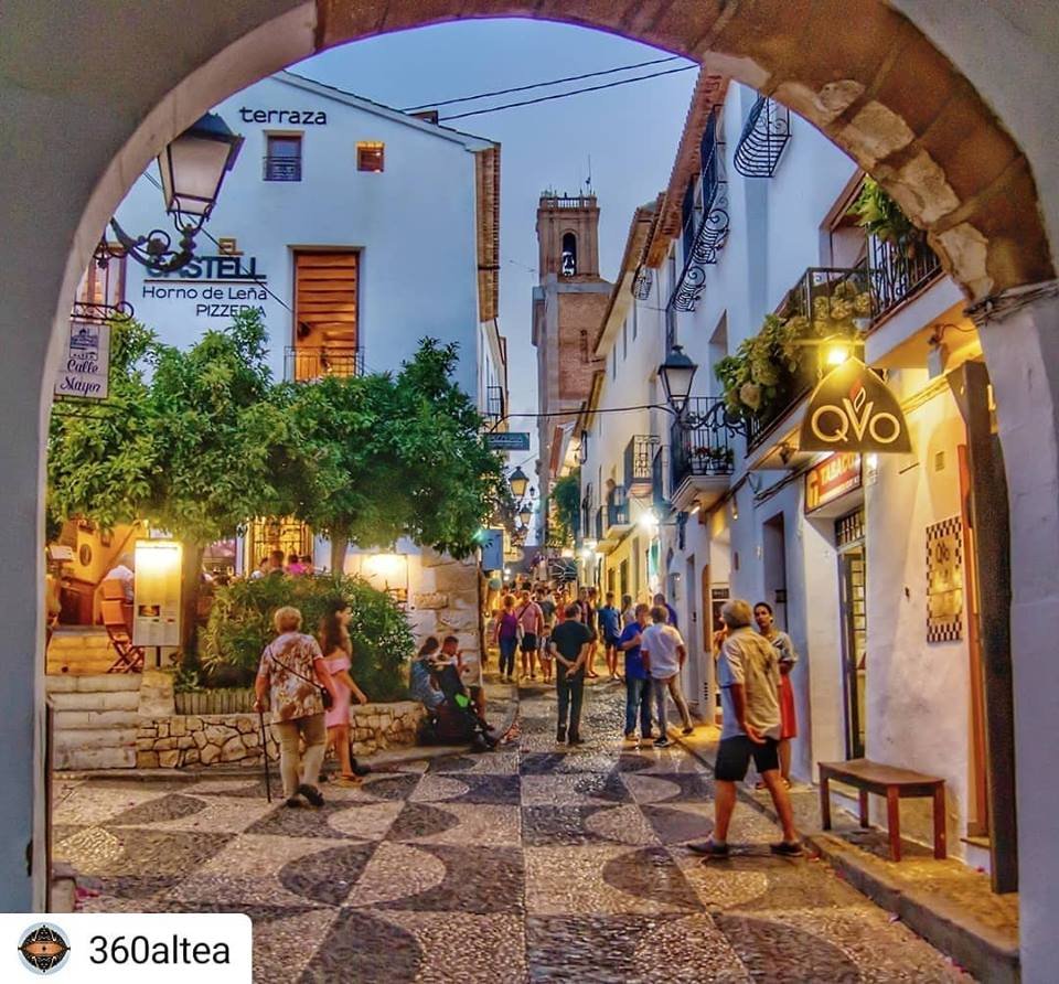 En travel gate i en liten by med mennesker som går og prater på kvelden. Det er bygninger med butikkutsalg og blomster på balkongene. Gatelyktene er tent.