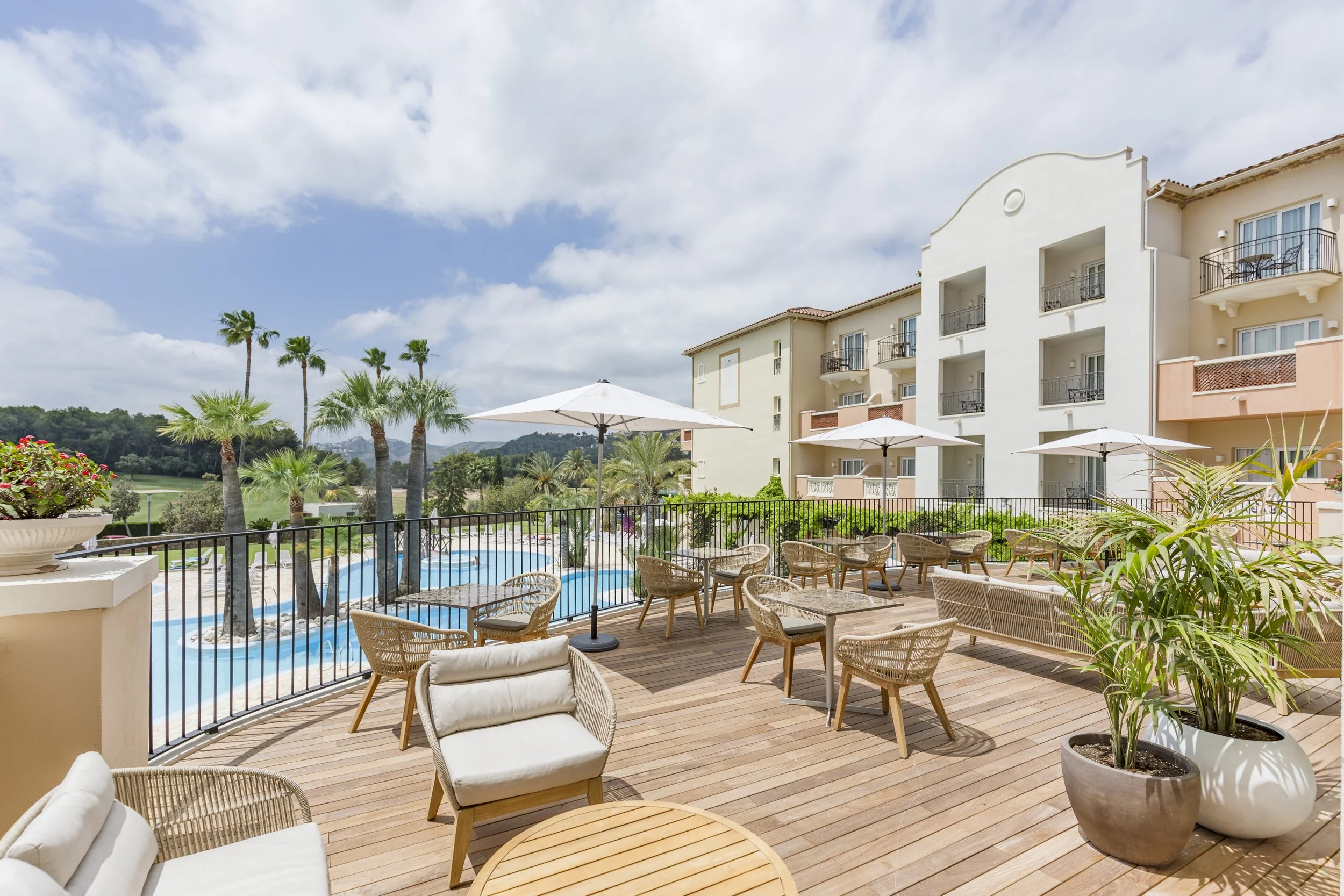Utsikt over en skyggefull utendørs terrasse med stoler, bord og parasoller, omgitt av palmer og et svømmebasseng, i bakgrunnen et hvitt hotell under en skyet himmel.