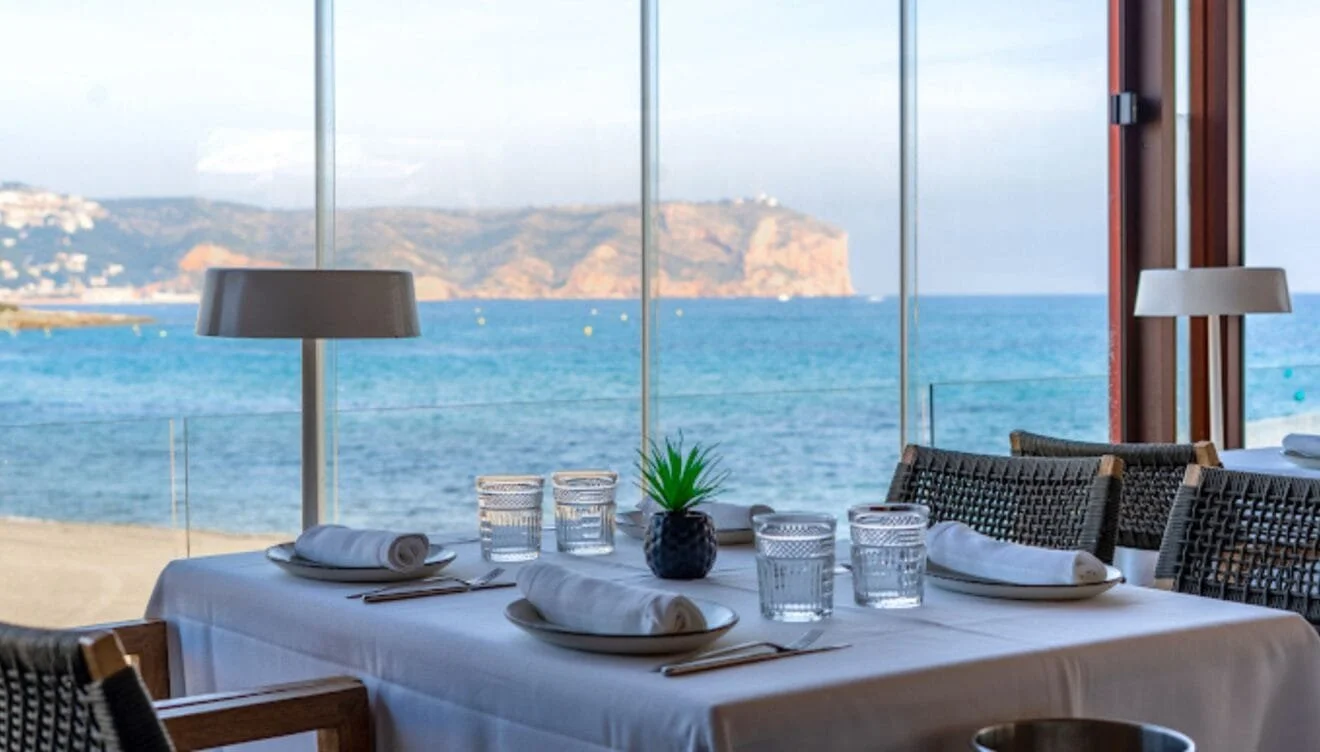 A set table in front of a panoramic window overlooking the Jávea bay with a mountain in the background at La Perla de Jávea restaurant