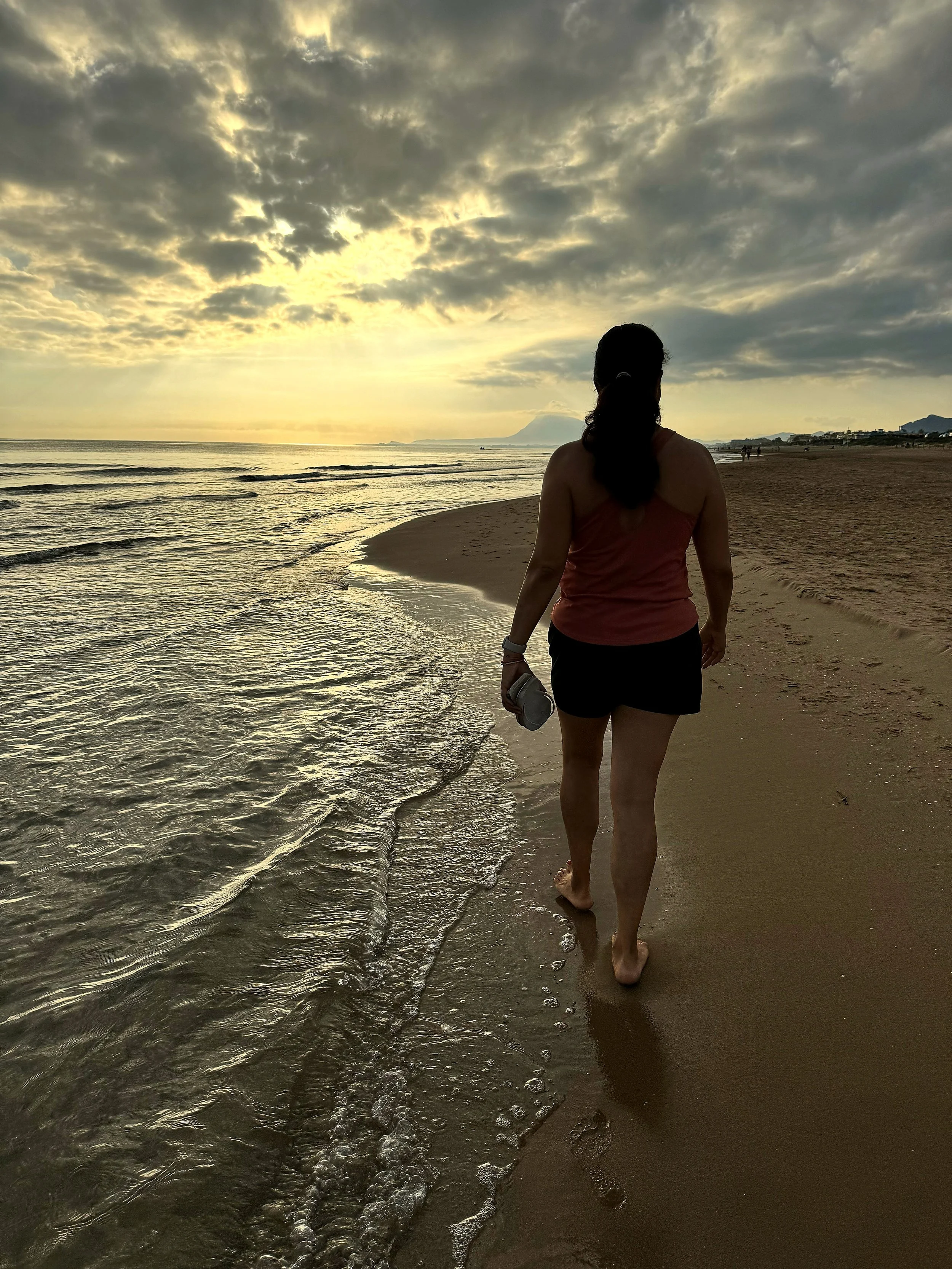 Una mujer camina por la playa al atardecer, con el agua chapoteando contra sus pies y el cielo lleno de nubes