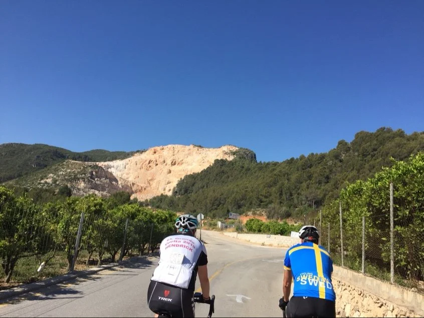 Two men cycling on a paved road through orange groves with forests and a quarry in the background