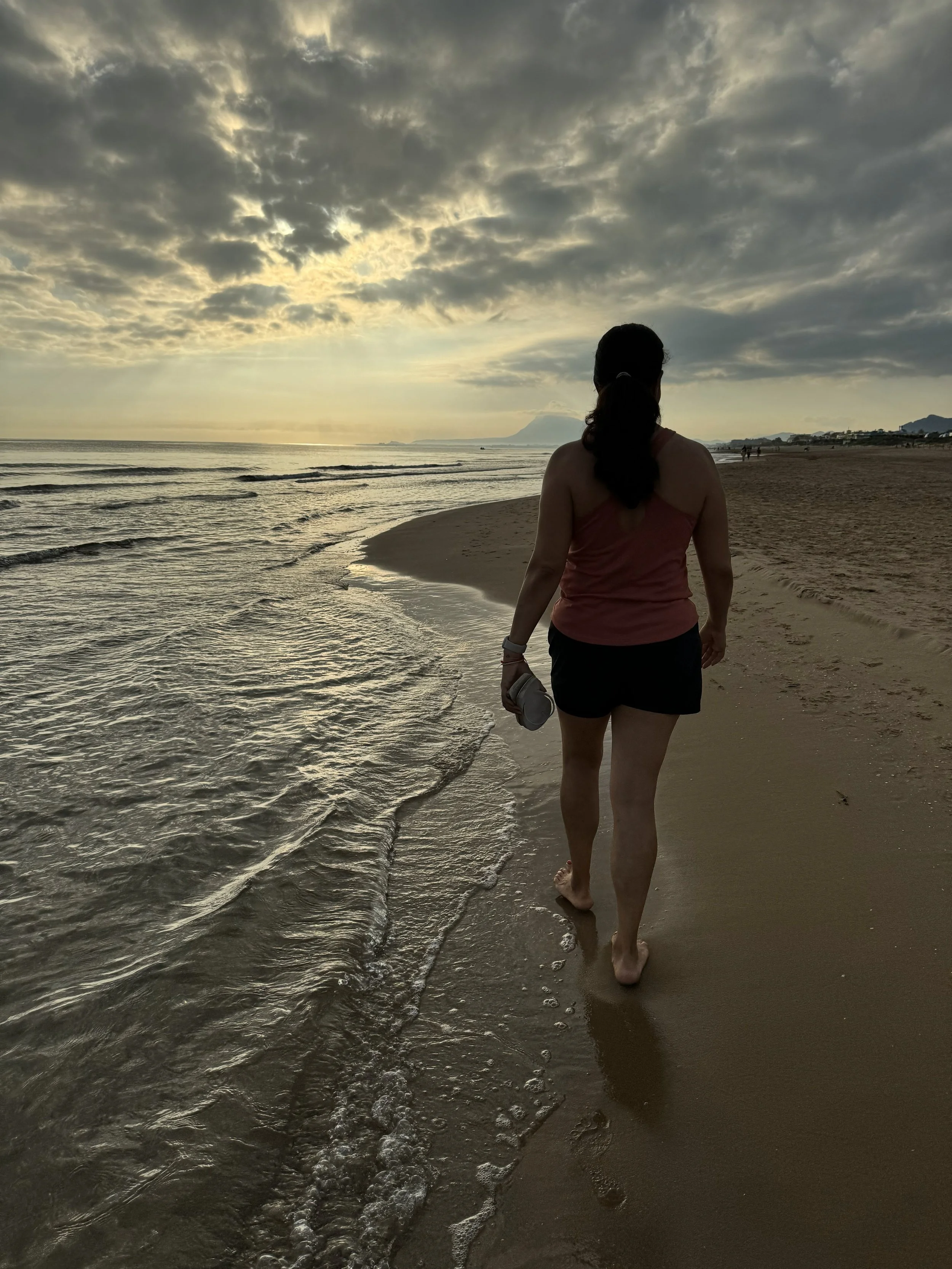 En kvinne går langs en sandstrand mot solnedgangen, små bølger ruller mot stranden med havet på den ene siden og fjell i bakgrunnen.