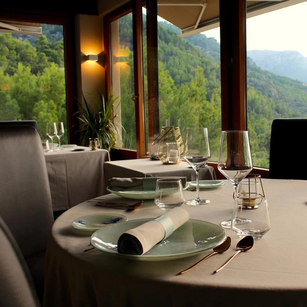 A set table in front of a panoramic window with a forest in the background at restaurant El Carreter in La Xara