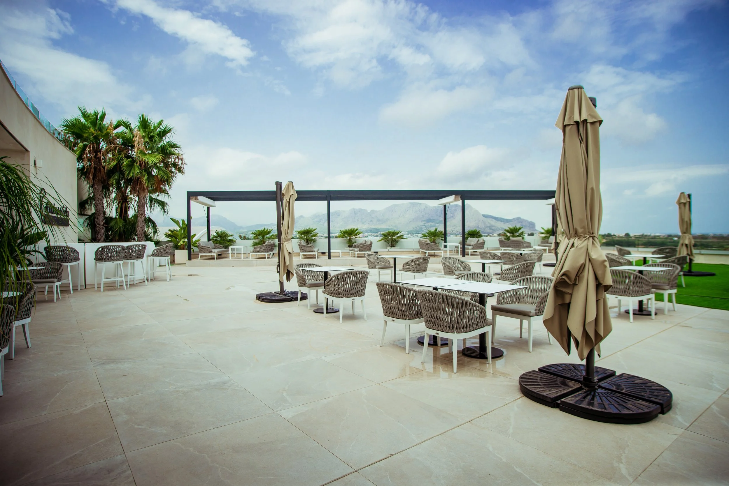 Utendørs terrasse med utemøbler, parasoller, palmer og fjell i bakgrunnen under en skyet himmel.