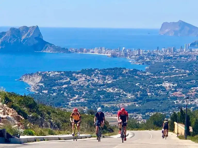 Three cyclists ascending a mountain with a city and mountains in the background