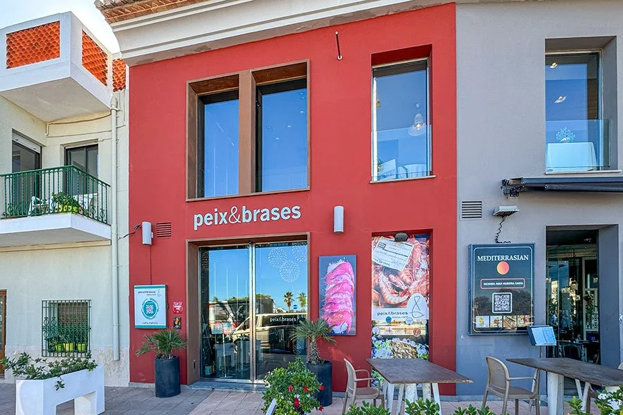 The red facade of Peix & Brases restaurant in Dénia with green plants and an outdoor table in the foreground