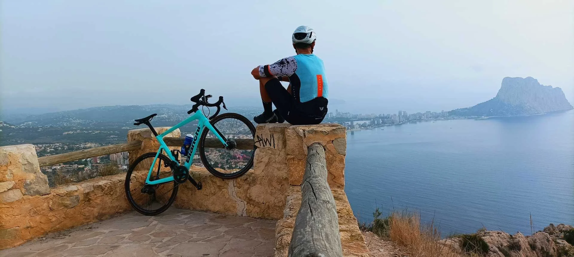 A man sitting on a stone fence beside his bicycle looking down towards a city and the sea in the distance