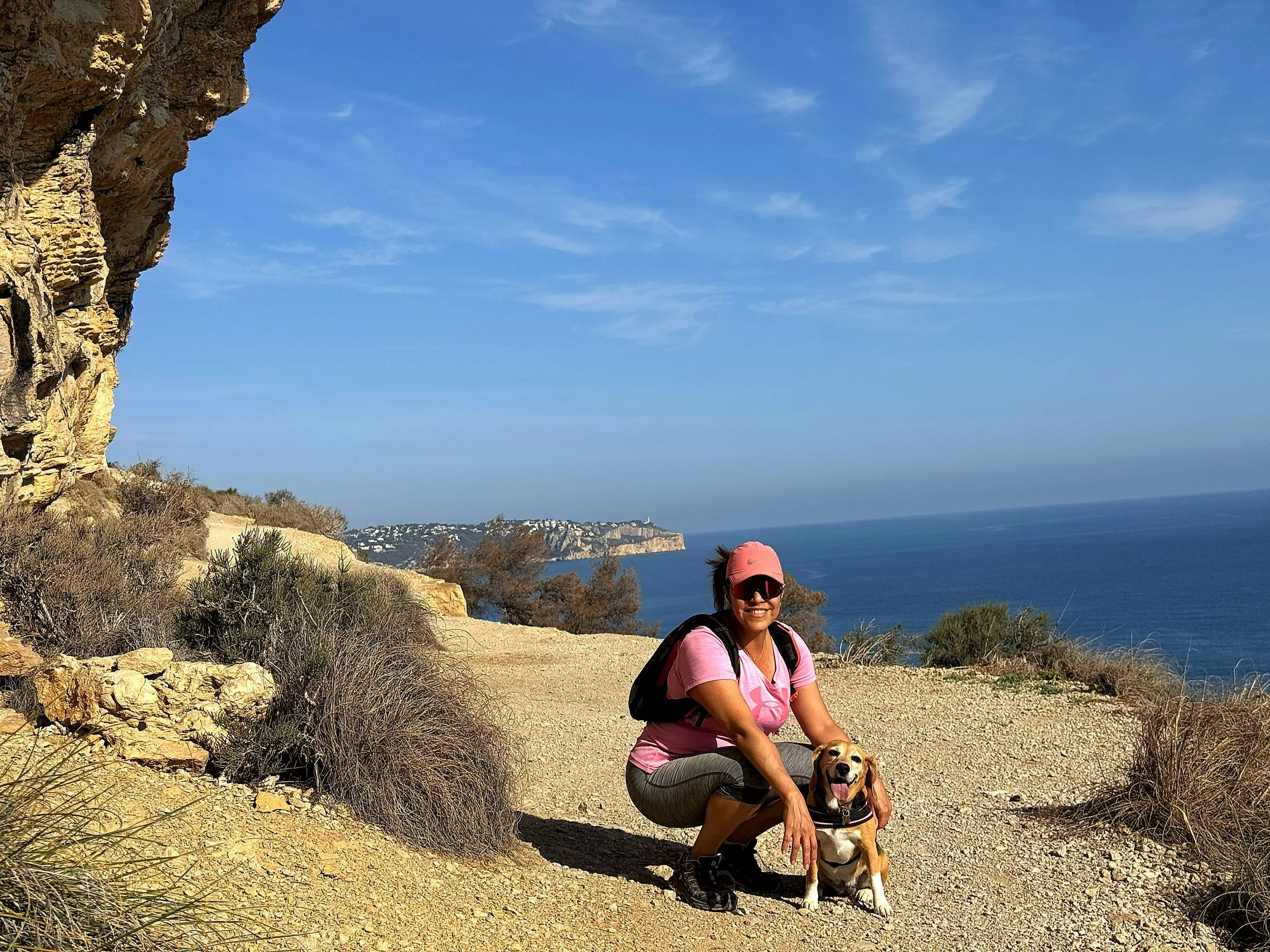 Mujer con gorra rosa, gafas de sol y camiseta rosa arrodillada junto a un perro en un sendero de grava, con el mar y acantilados al fondo