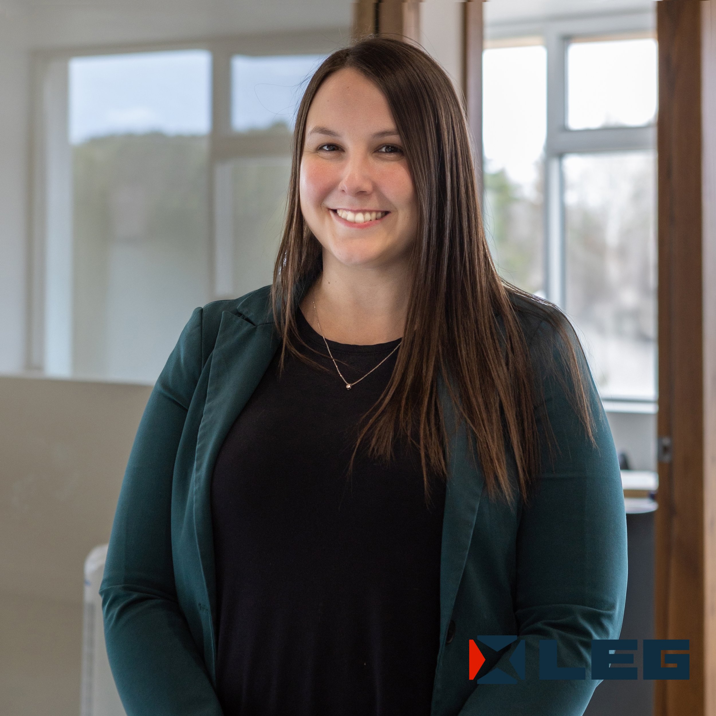 Une femme souriante avec les cheveux bruns longs, portant un blazer vert foncé et un t-shirt noir, dans un espace intérieur avec de grandes fenêtres