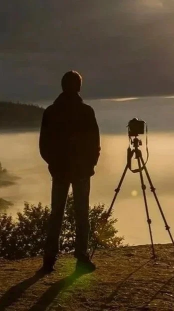 Personne debout près d'un appareil photo sur un trépied, regardant un paysage avec de l'eau et la forêt au coucher du soleil.