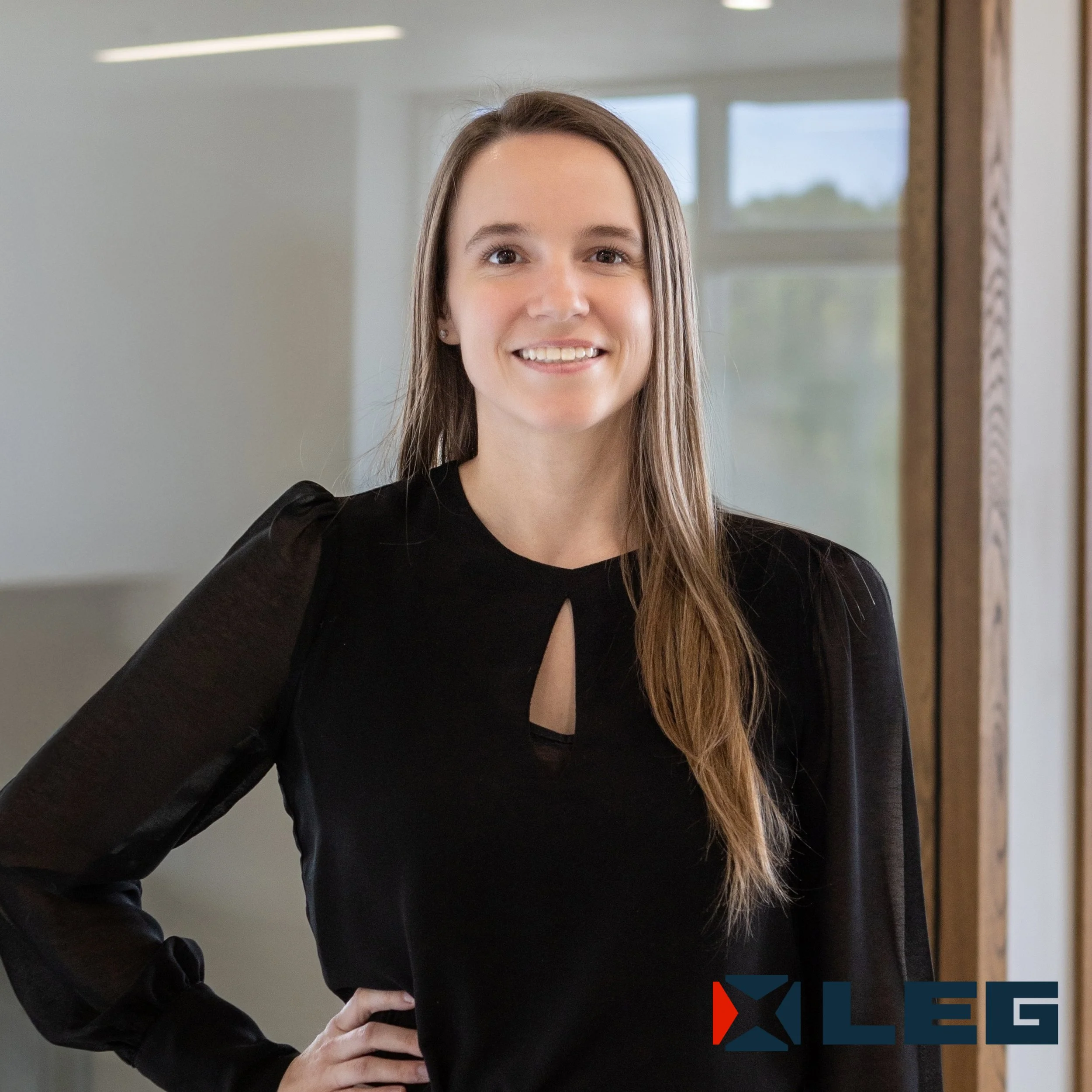 Jeune femme brune souriante portant un haut noir avec des manches longues, posant en intérieur près d'une fenêtre.