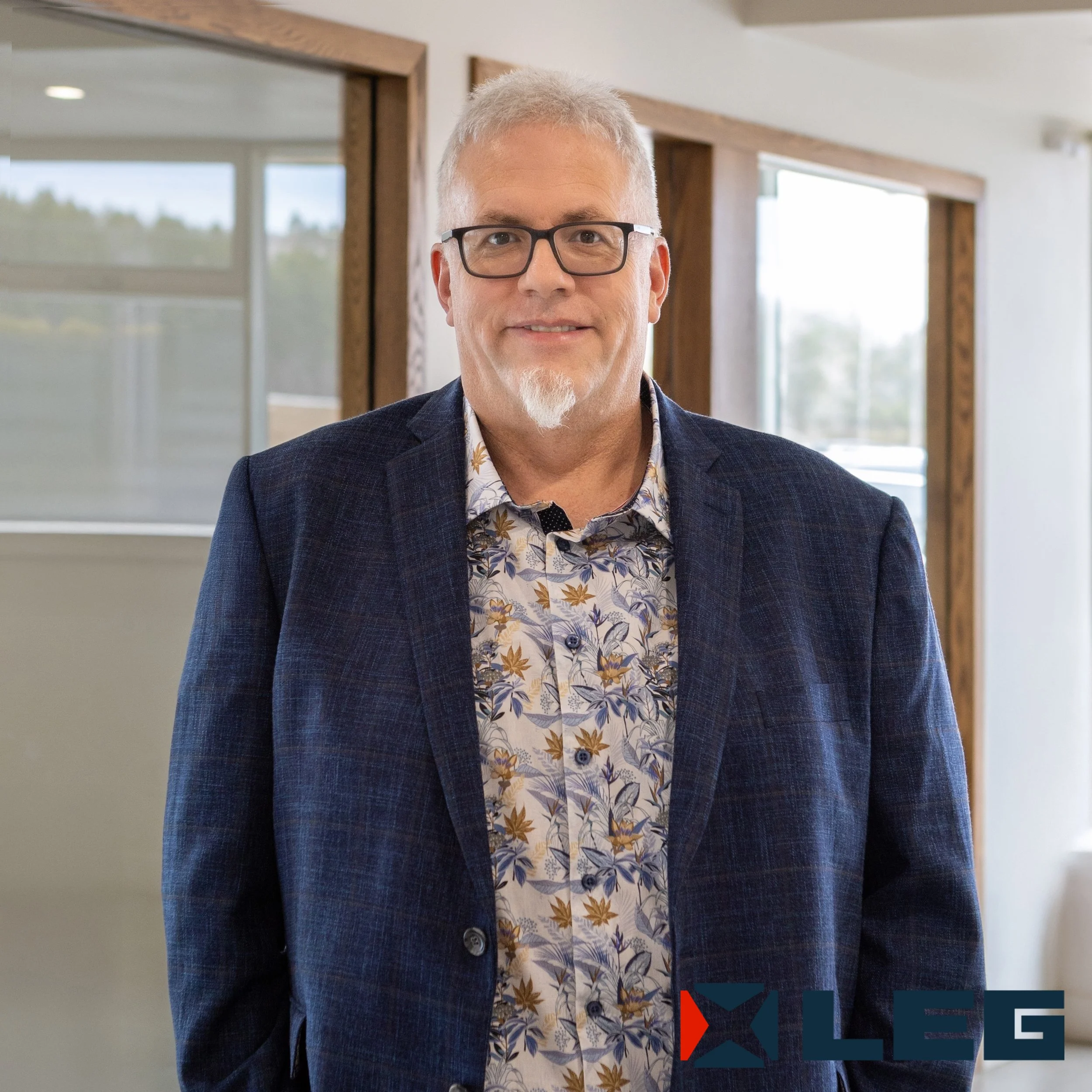 Un homme portant des lunettes, une chemise à motifs floraux et une veste en costume, posant à l'intérieur d'un bâtiment moderne avec de grandes fenêtres.