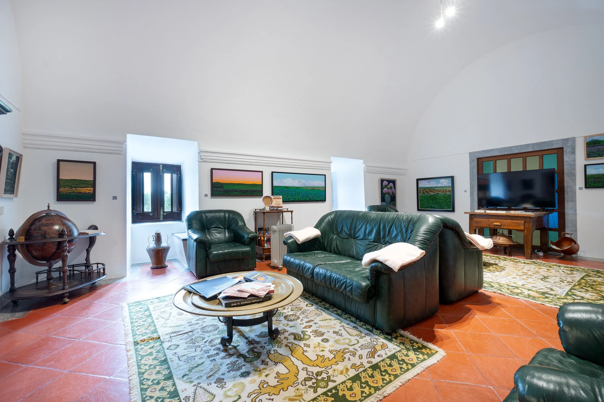 Sala de estar com sofá de couro verde, retratos de paisagens na parede, televisão em móvel de madeira, tapete e piso de ladrilho, janelas com cortinas brancas.