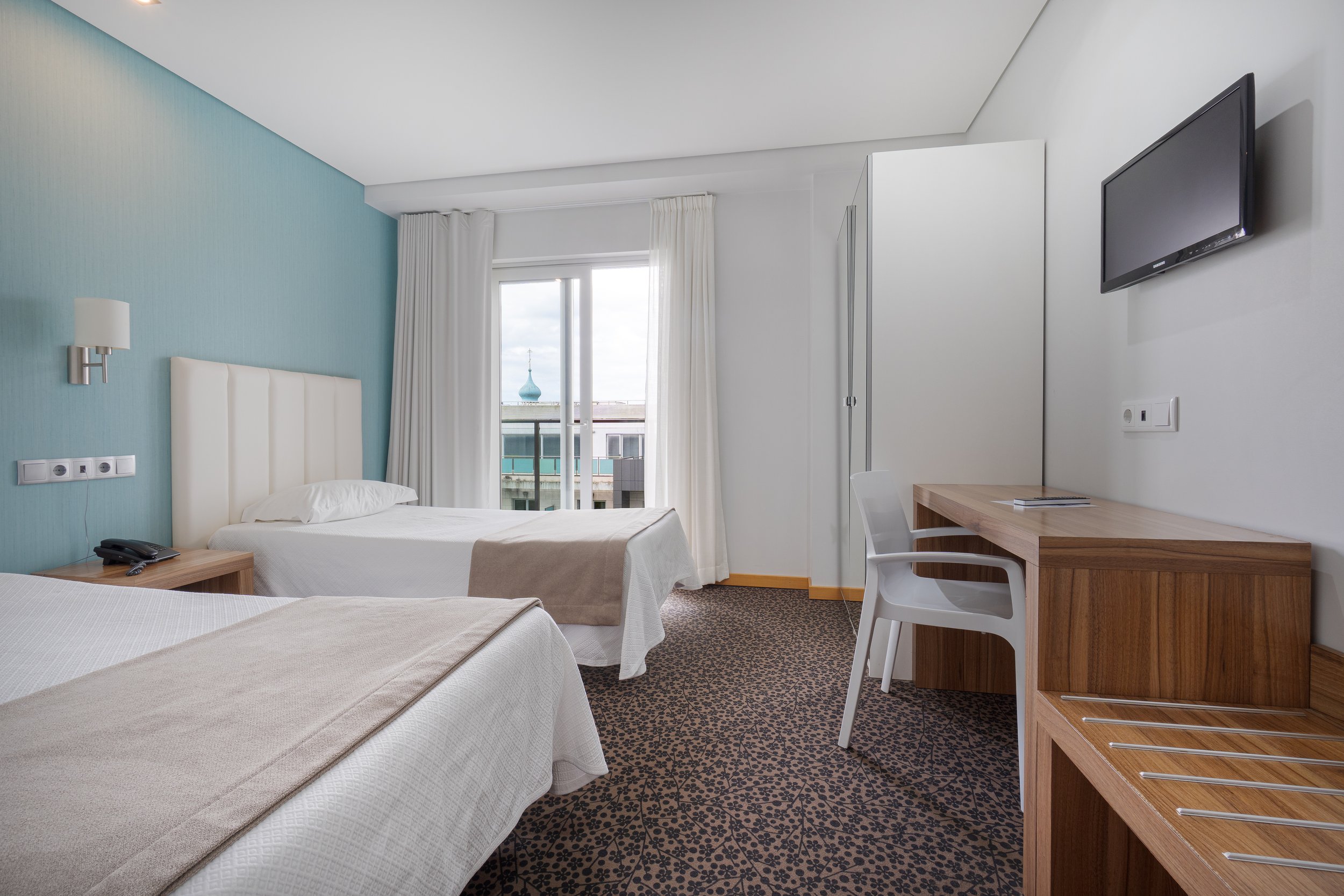 Quarto de hotel com duas camas, uma com lençol branco e outra com lençol bege, vista para uma varanda com porta de vidro, televisão na parede, mesa de trabalho com cadeira branca, armário branco e piso de carpete com padrão escuro.