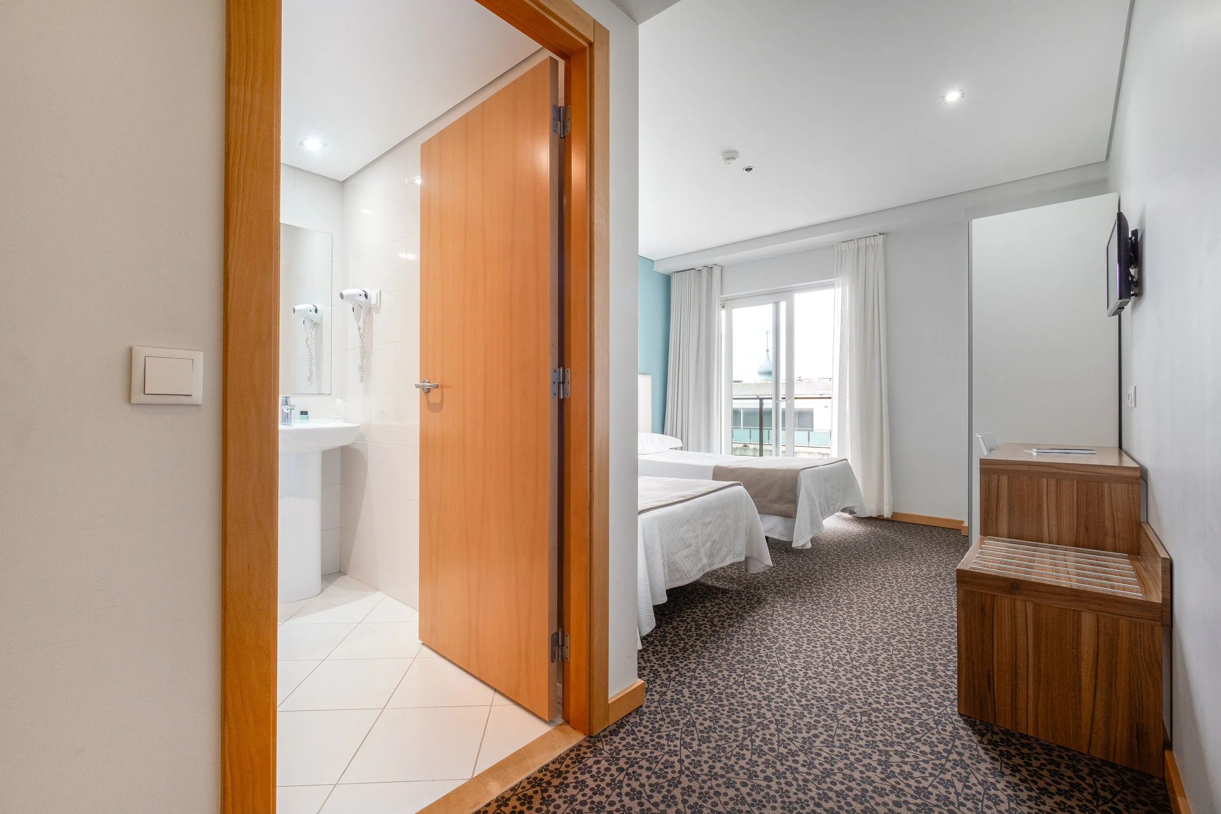 Quarto de hotel com duas camas, televisão na parede, porta para banheiro, janela com cortinas e vista para edifícios.