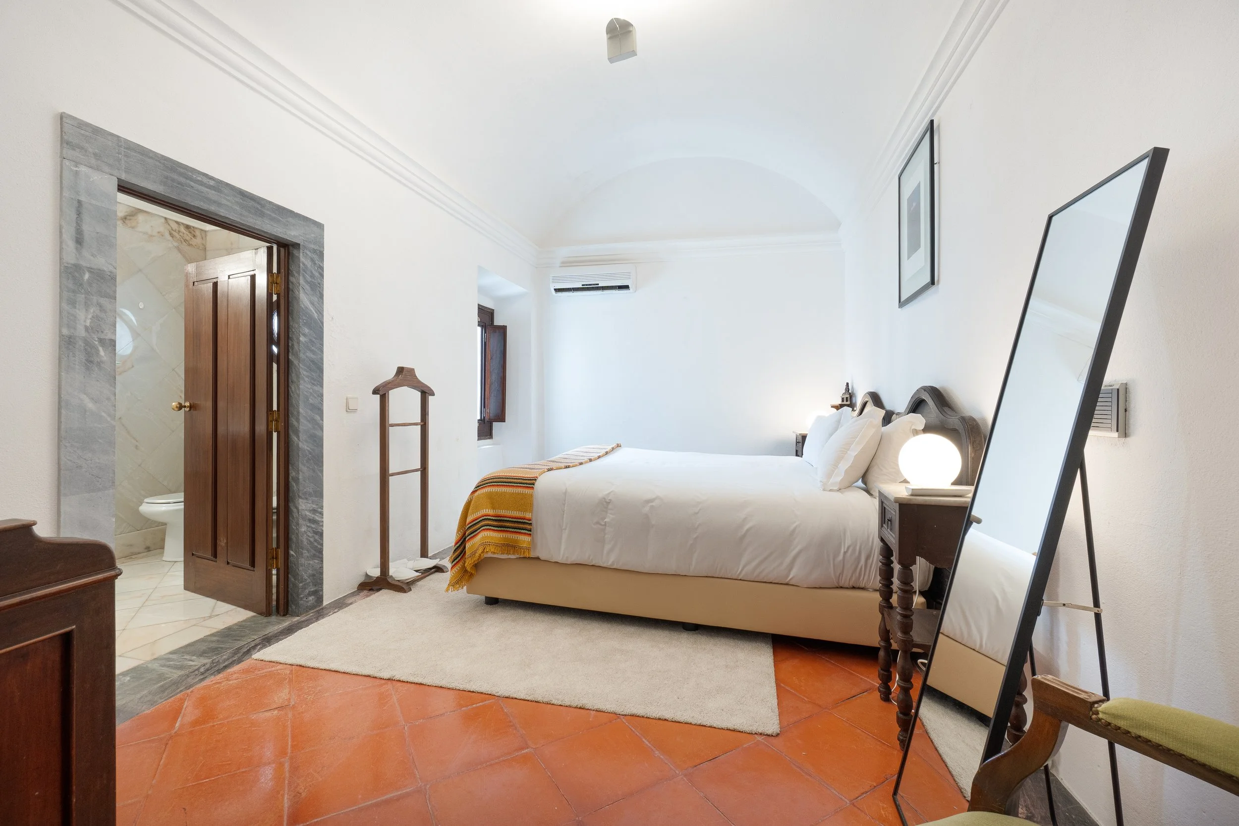Quarto de hotel com cama de casal, espelho de corpo inteiro em suporte, abajur na mesa de cabeceira, parede branca, piso de cerâmica alaranjada, e banheiro visível ao lado com porta de madeira aberta.