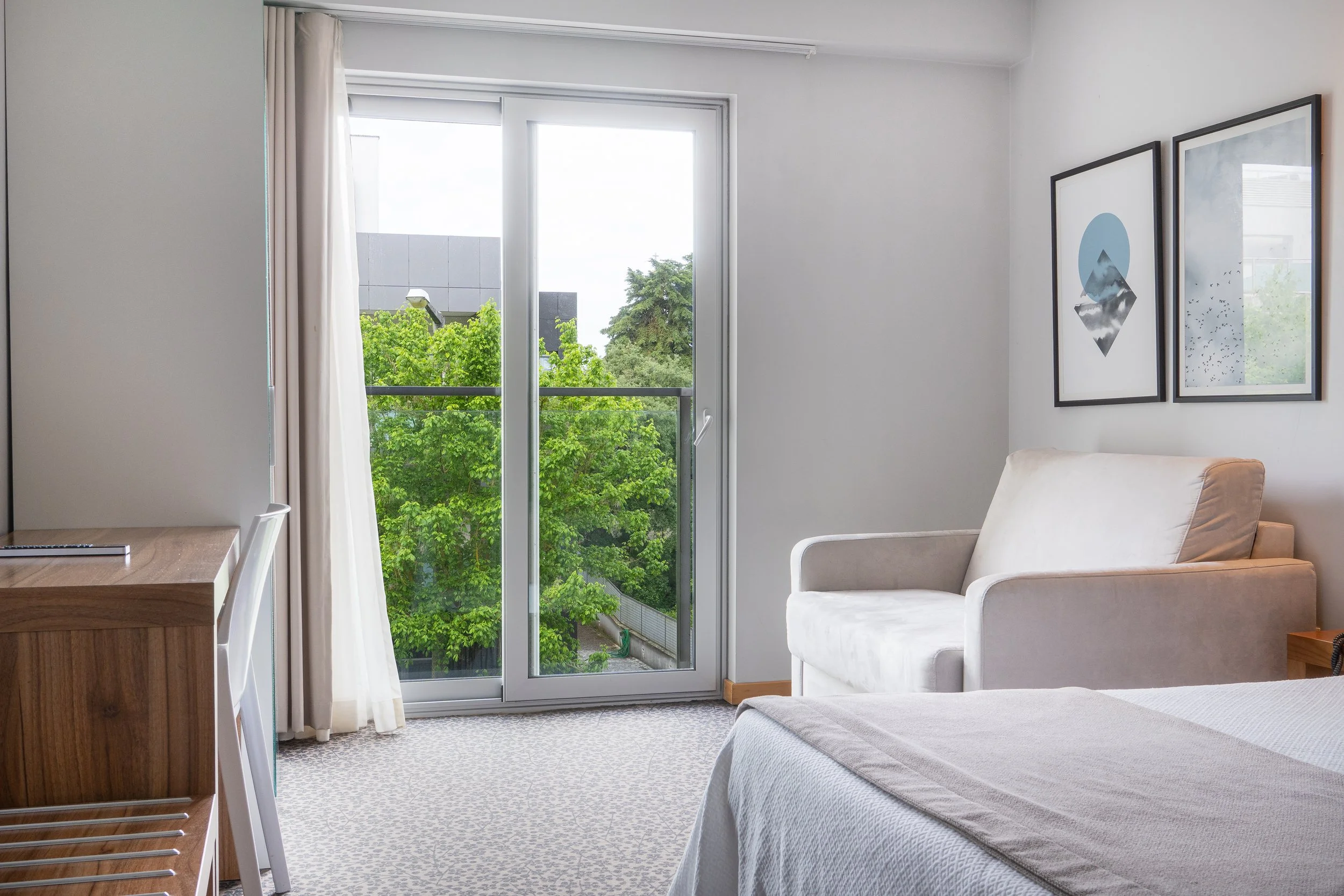 Quarto de hotel com cama, sofá branco, janela de vidro com cortinas brancas, árvores visíveis do lado de fora e quadros decorativos na parede.