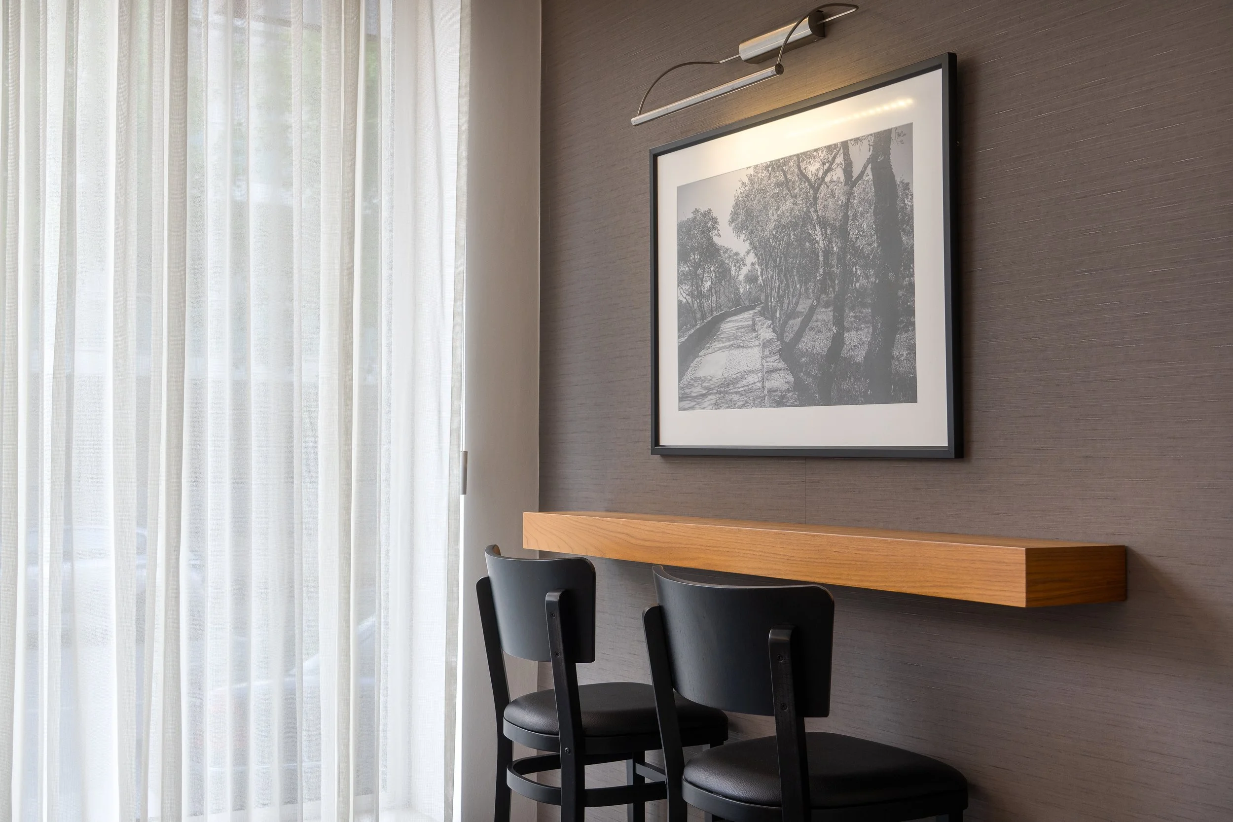 Sala com parede de textura cinza, uma moldura preta com fotografia em tons de cinza de uma estrada com árvores, uma prateleira de madeira, duas cadeiras pretas com assento de couro, cortinas brancas que deixam a luz entrar.