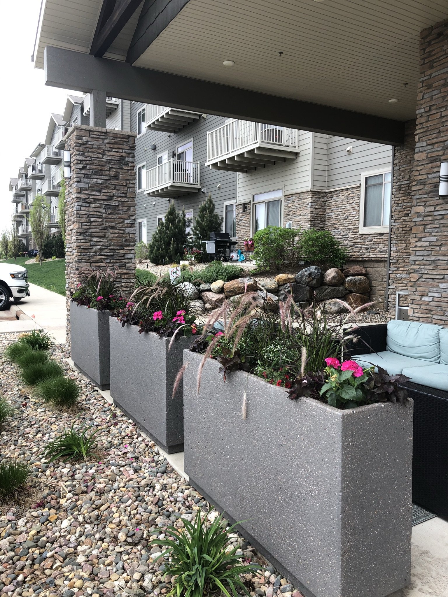 Apartment building patio area with large gray concrete planters filled with pink and purple flowers, ornamental grasses, and green shrubs, with some small green plants and a gravel pathway in the foreground.