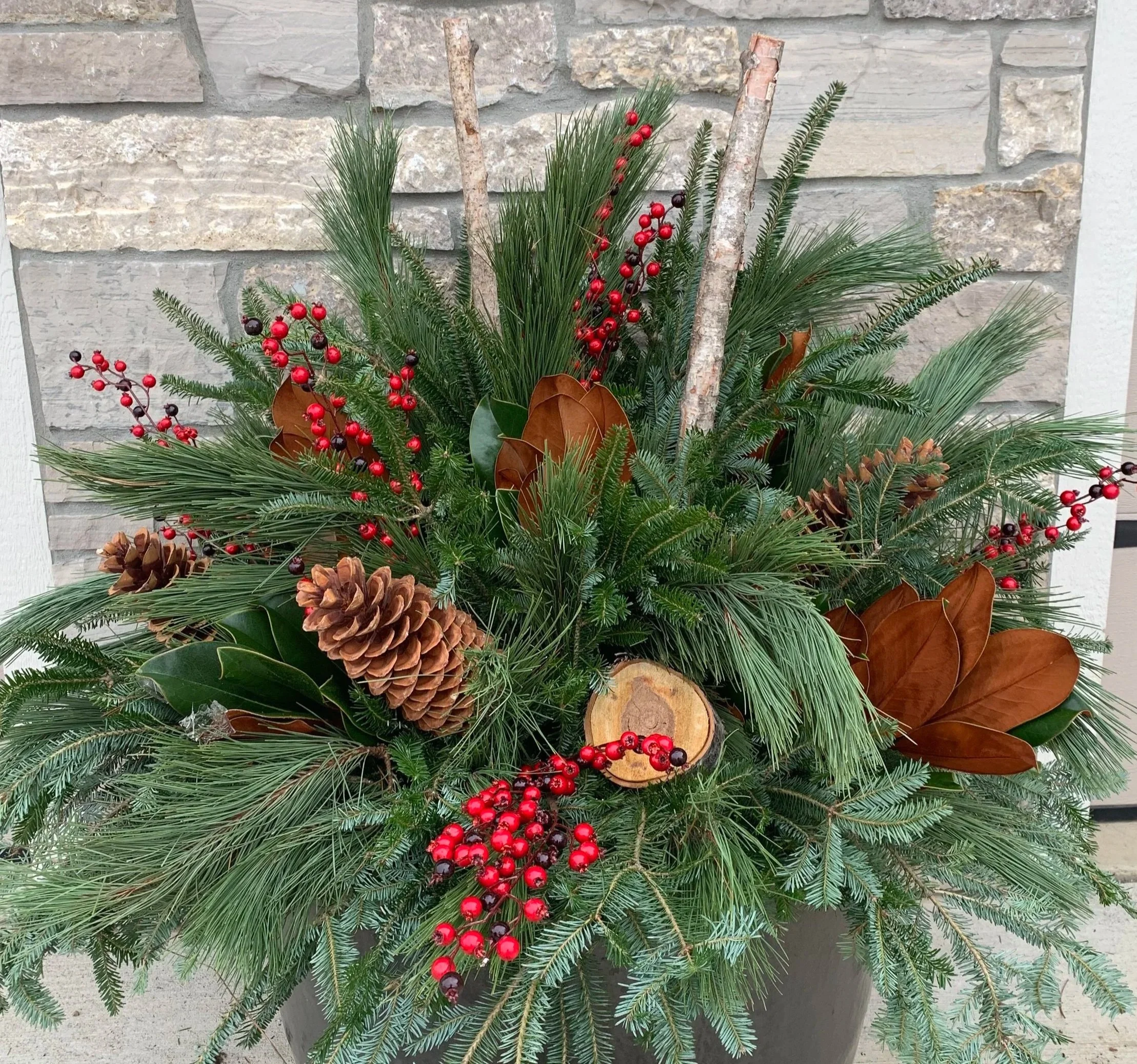 A festive holiday arrangement of evergreen branches, pinecones, red berries, and wooden accents in a container.